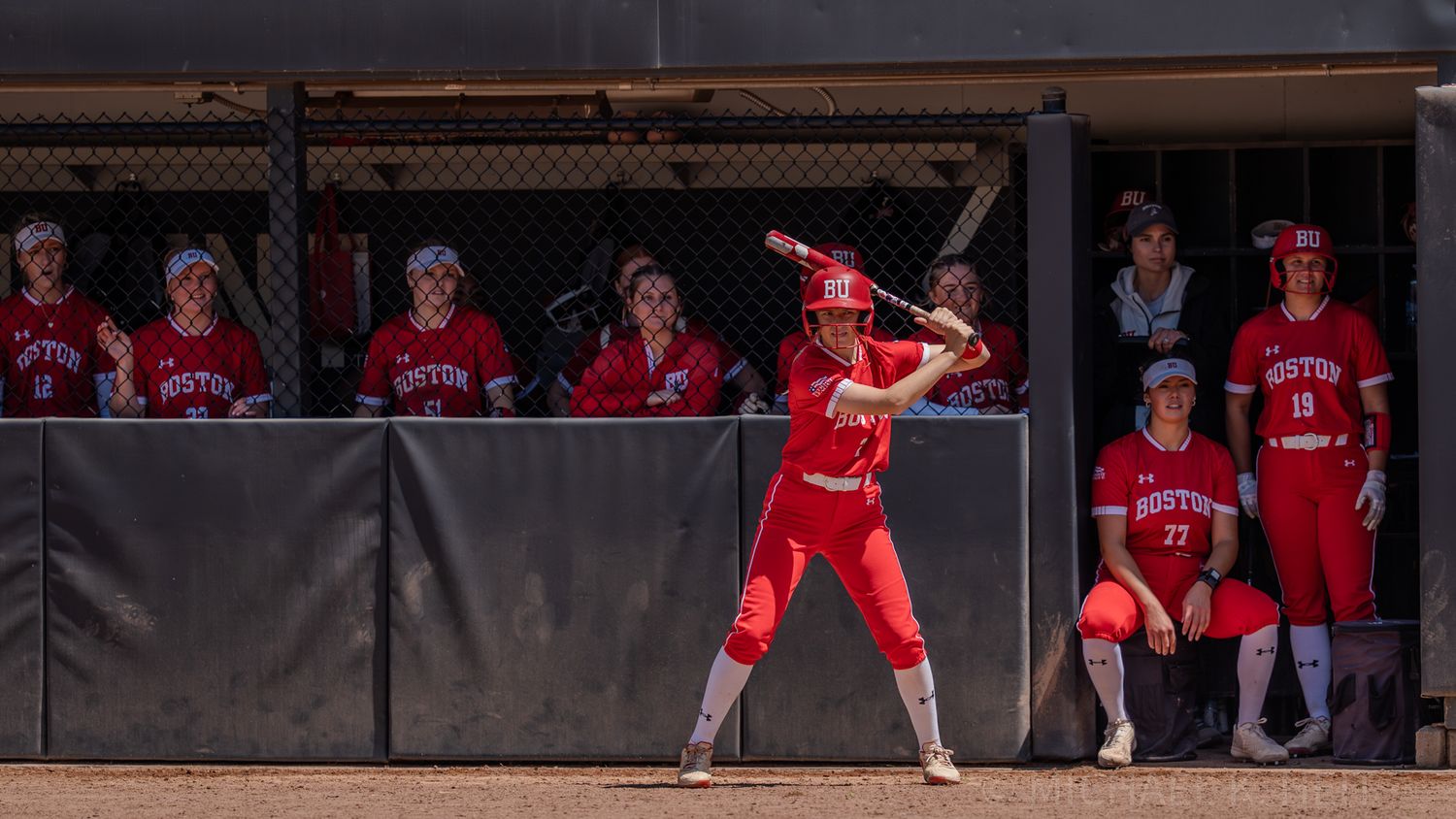 Softball player in red uniform at bat with teammates watching from dugout.