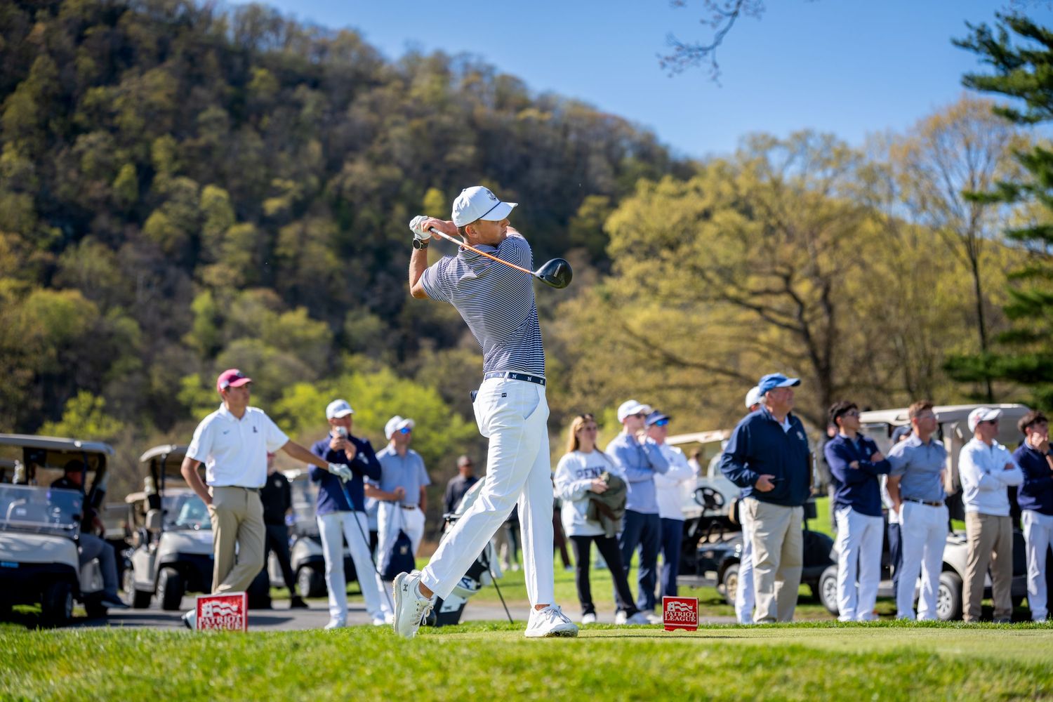 Golfer swinging a club on a lush green course with spectators watching on a sunny spring day.