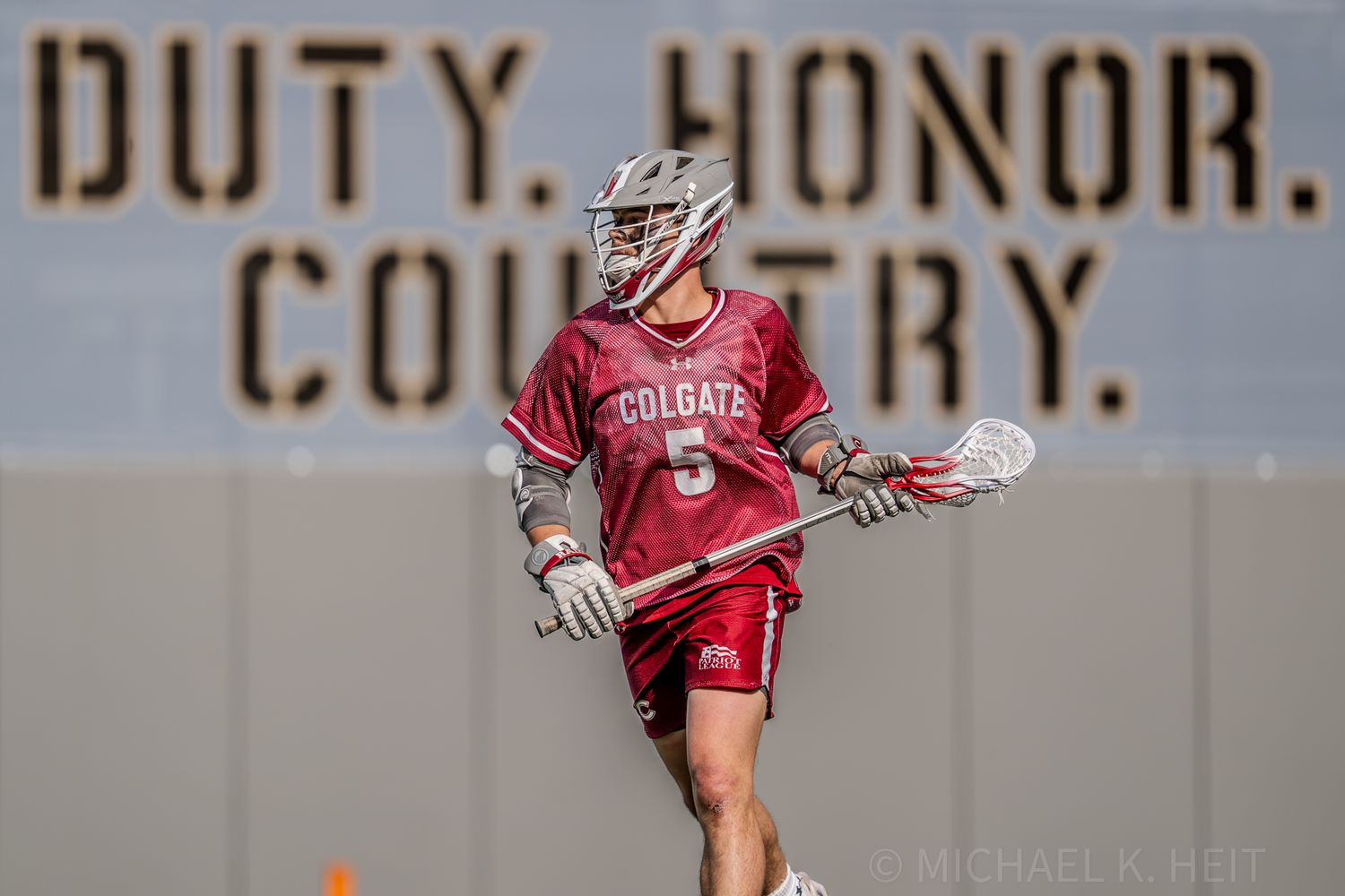 Lacrosse players compete intensely with text reading 'Duty Honor Country' visible in background.