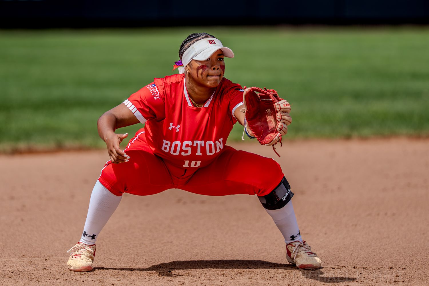 Softball player in red uniform crouches in defensive position on infield dirt.
