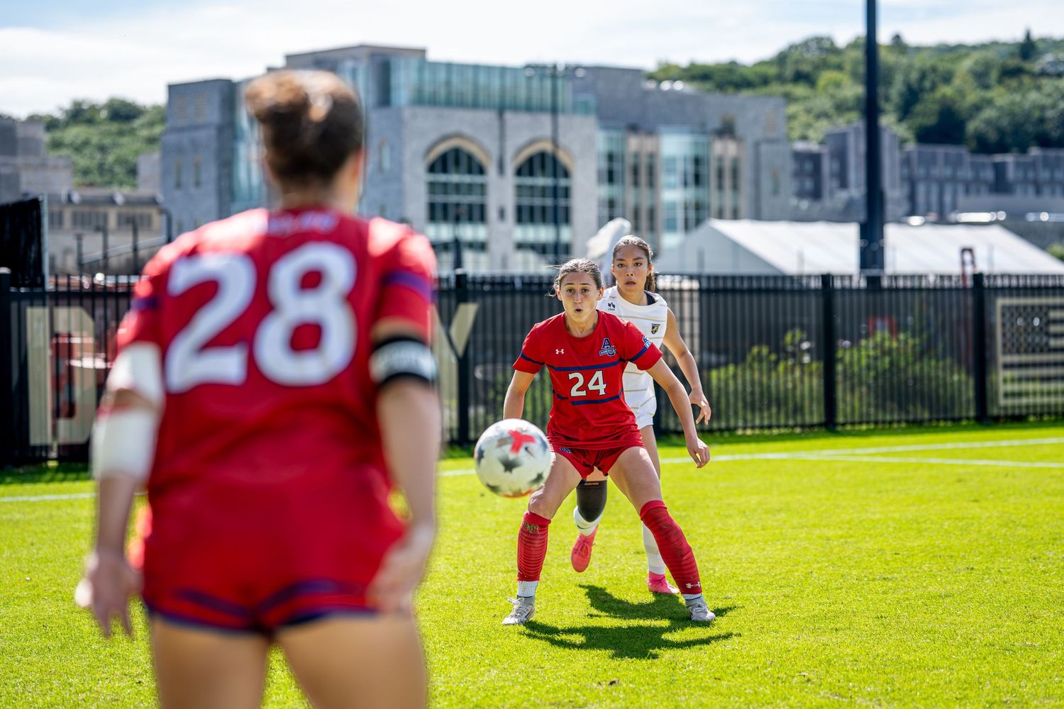 Two female soccer players in red uniforms compete for the ball on a green field during a sunny match.