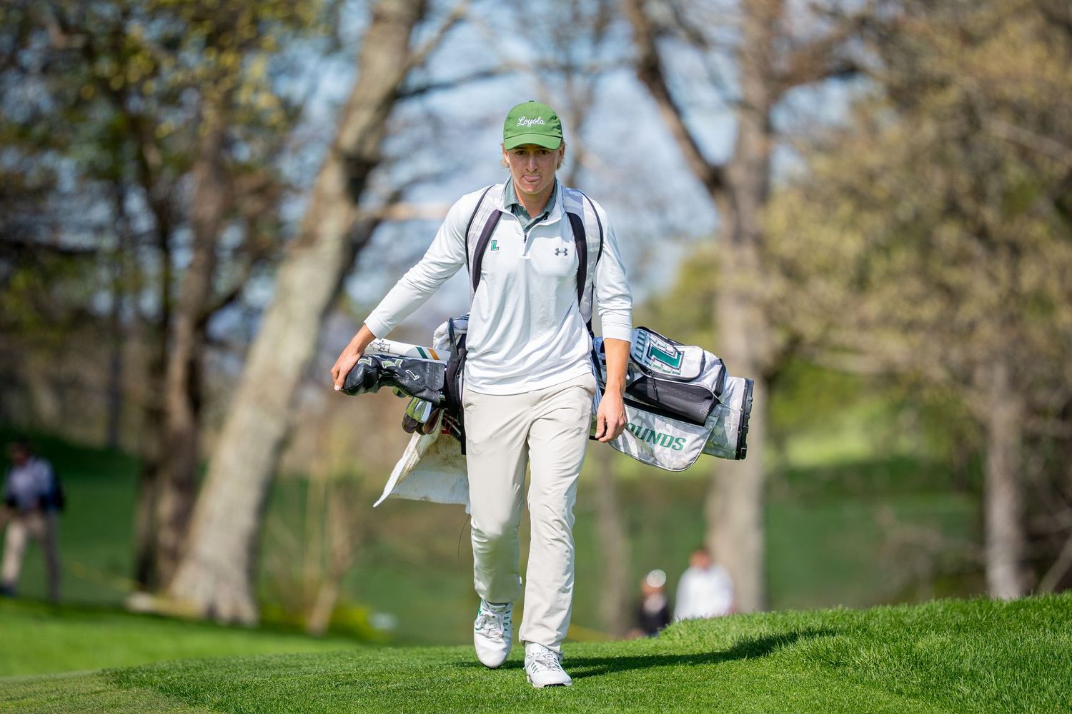 Golfer carrying a golf bag walks on a lush green fairway surrounded by trees on a sunny day.