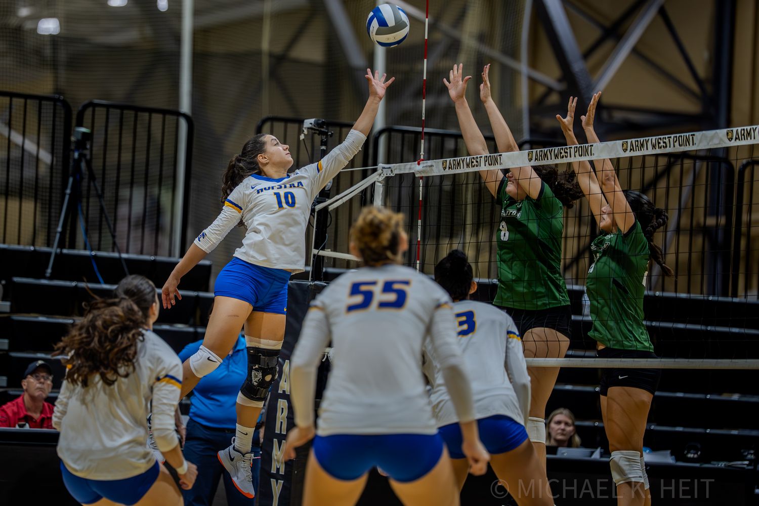 Indoor volleyball players compete at the net with one player attempting to block a spike.
