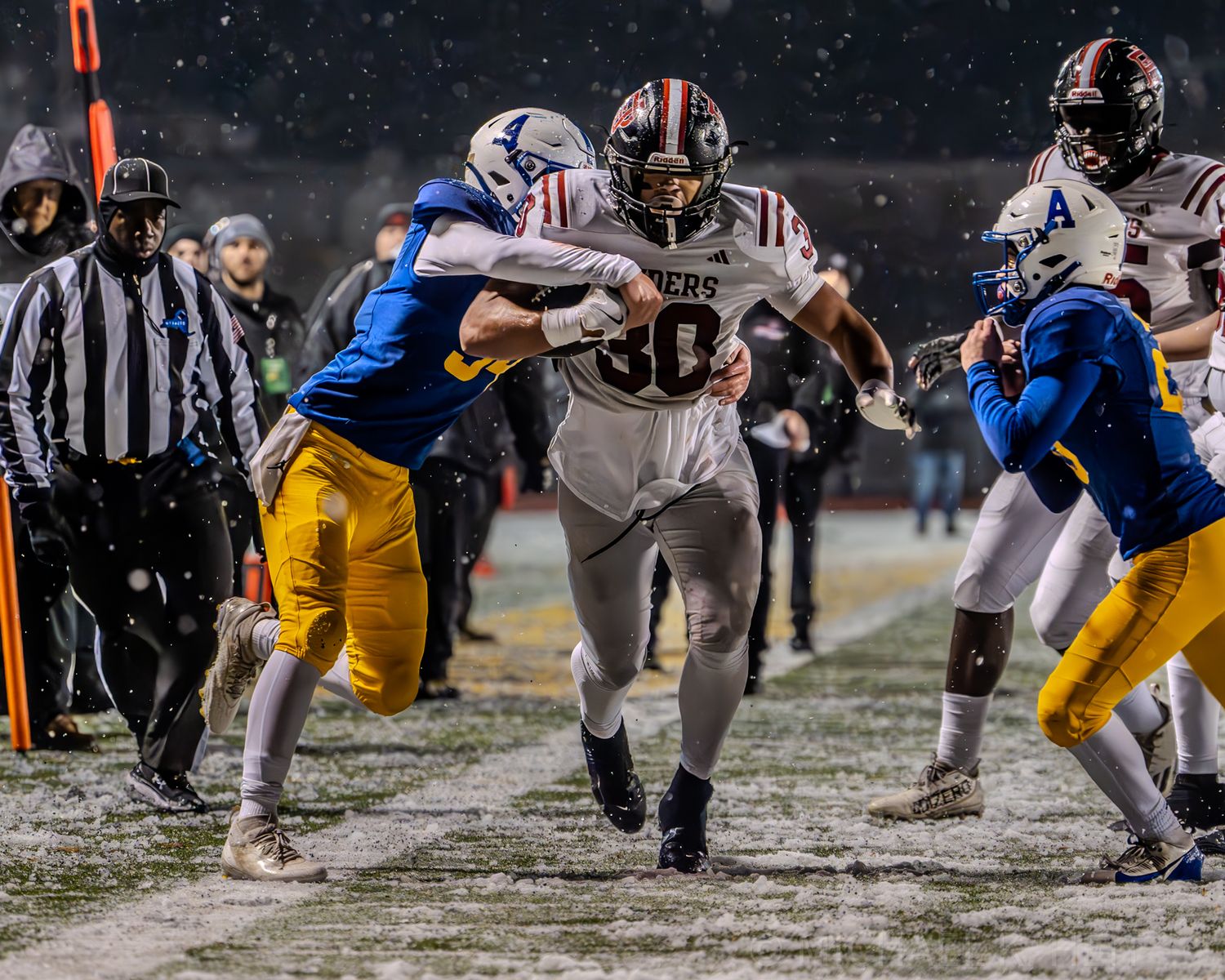 Football players battle in the snow during an intense ground play at night.