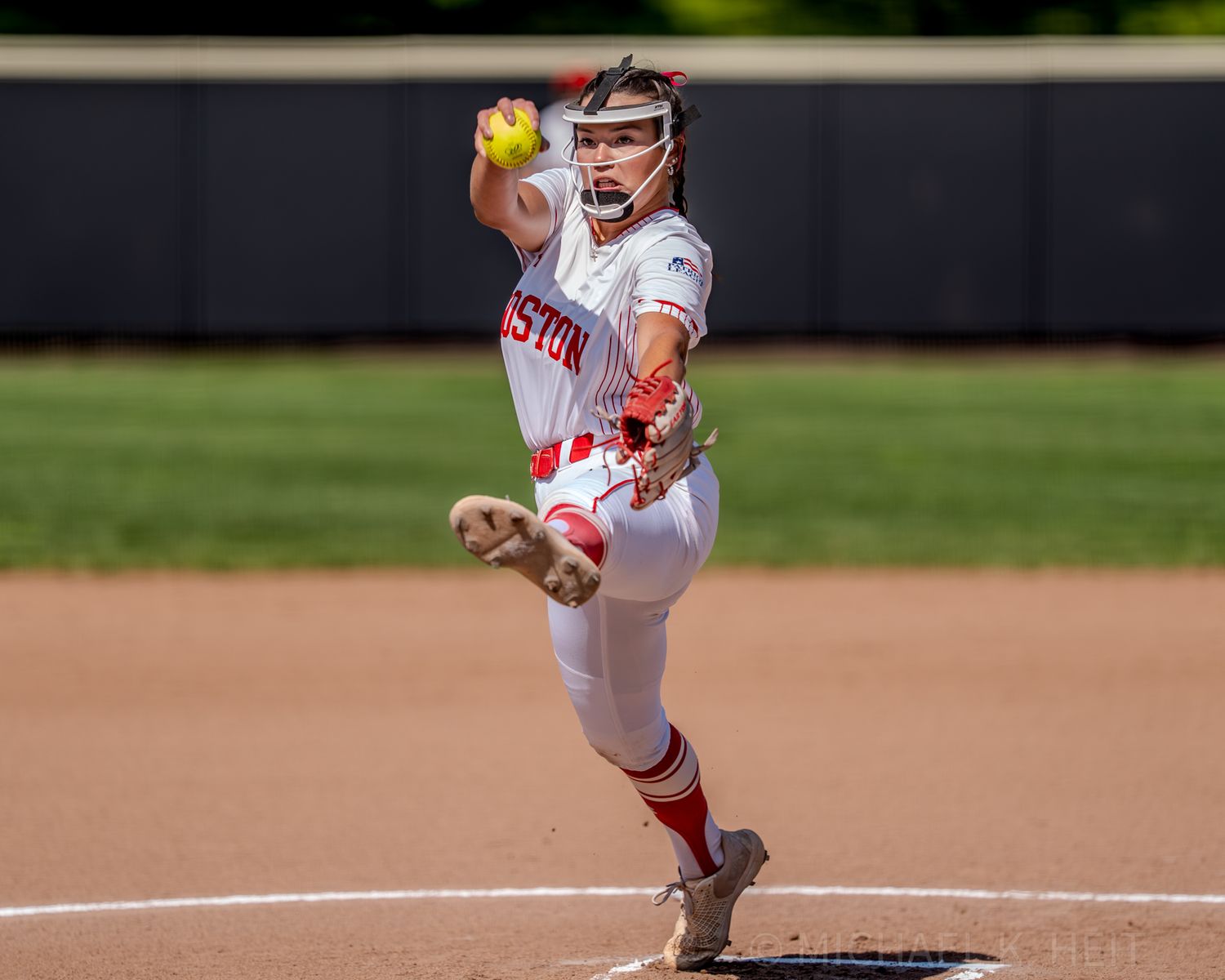 Softball pitcher in white and red Boston uniform winds up to throw during game.