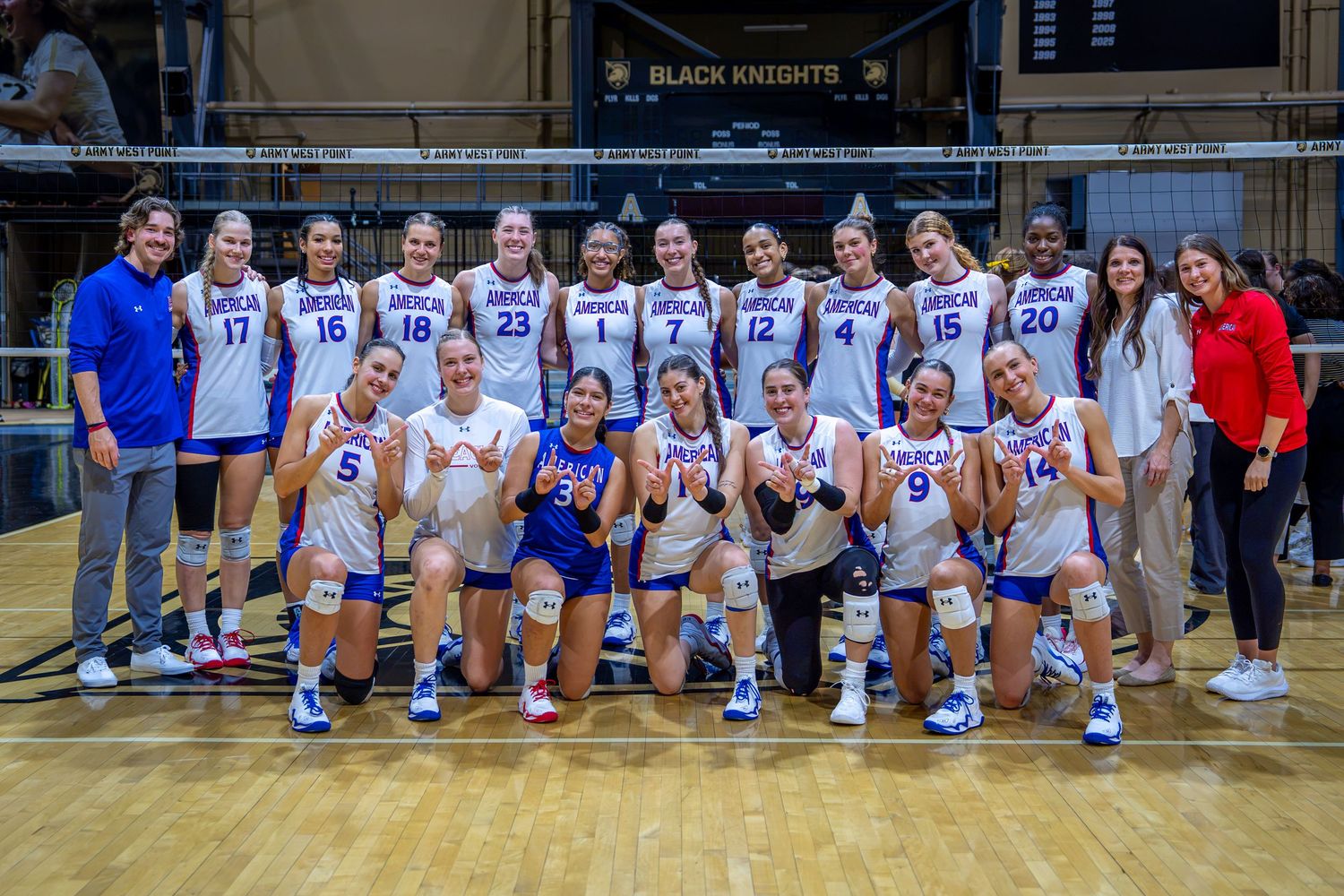Women's volleyball team poses together on court in blue and white uniforms under Black Knights banner.