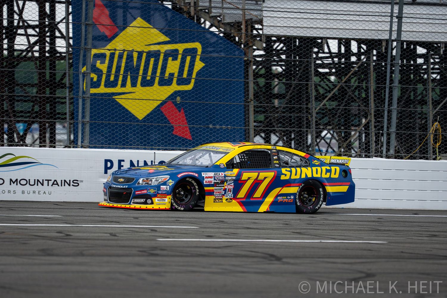 NASCAR #77 Sunoco sponsored race car speeding past Sunoco billboard at Pocono Raceway during a NASCAR Cup Series race.