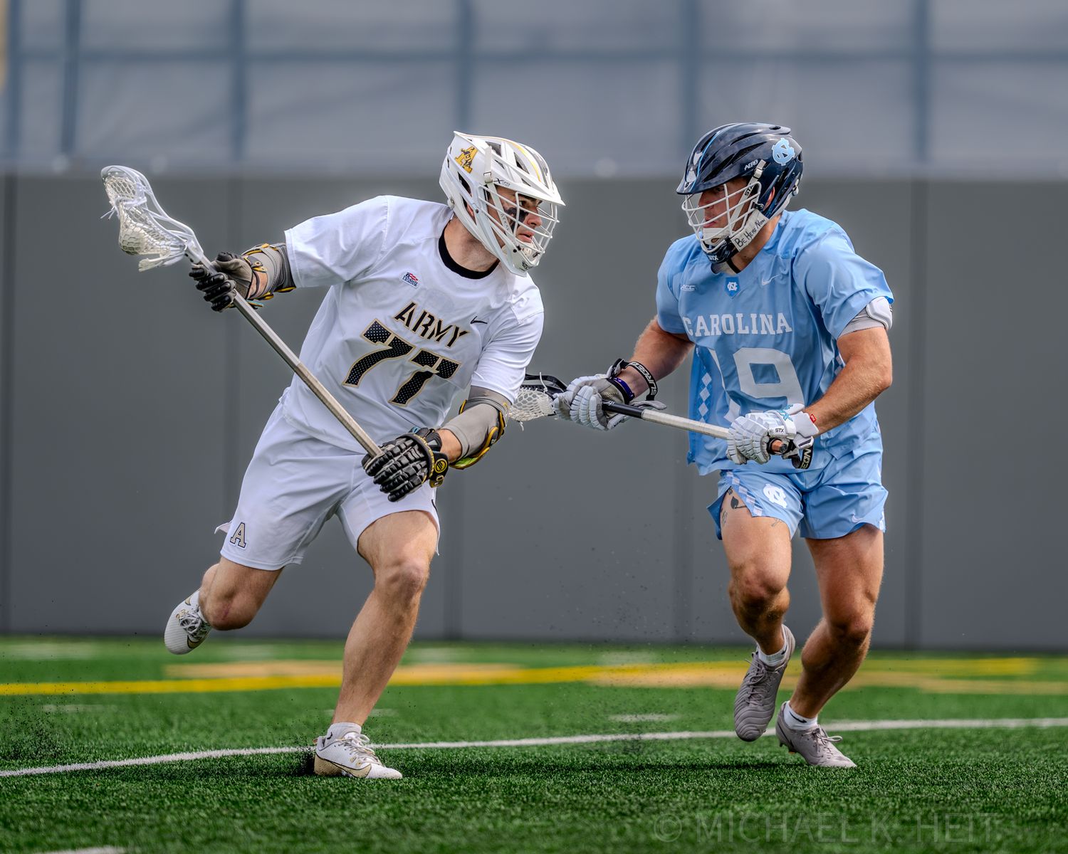 Lacrosse players in white and light blue uniforms compete during an intense match.
