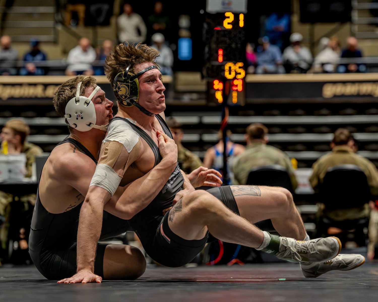 Two wrestlers compete on a mat during an indoor wrestling match, with a scoreboard visible in the background.