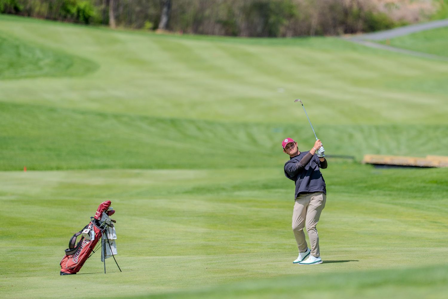 Golfer swinging a club on a lush green fairway with a red golf bag standing nearby on a sunny day.