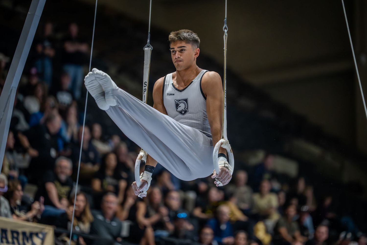 Male gymnast performing on still rings in a white uniform at an Army gymnastics competition before a packed arena.