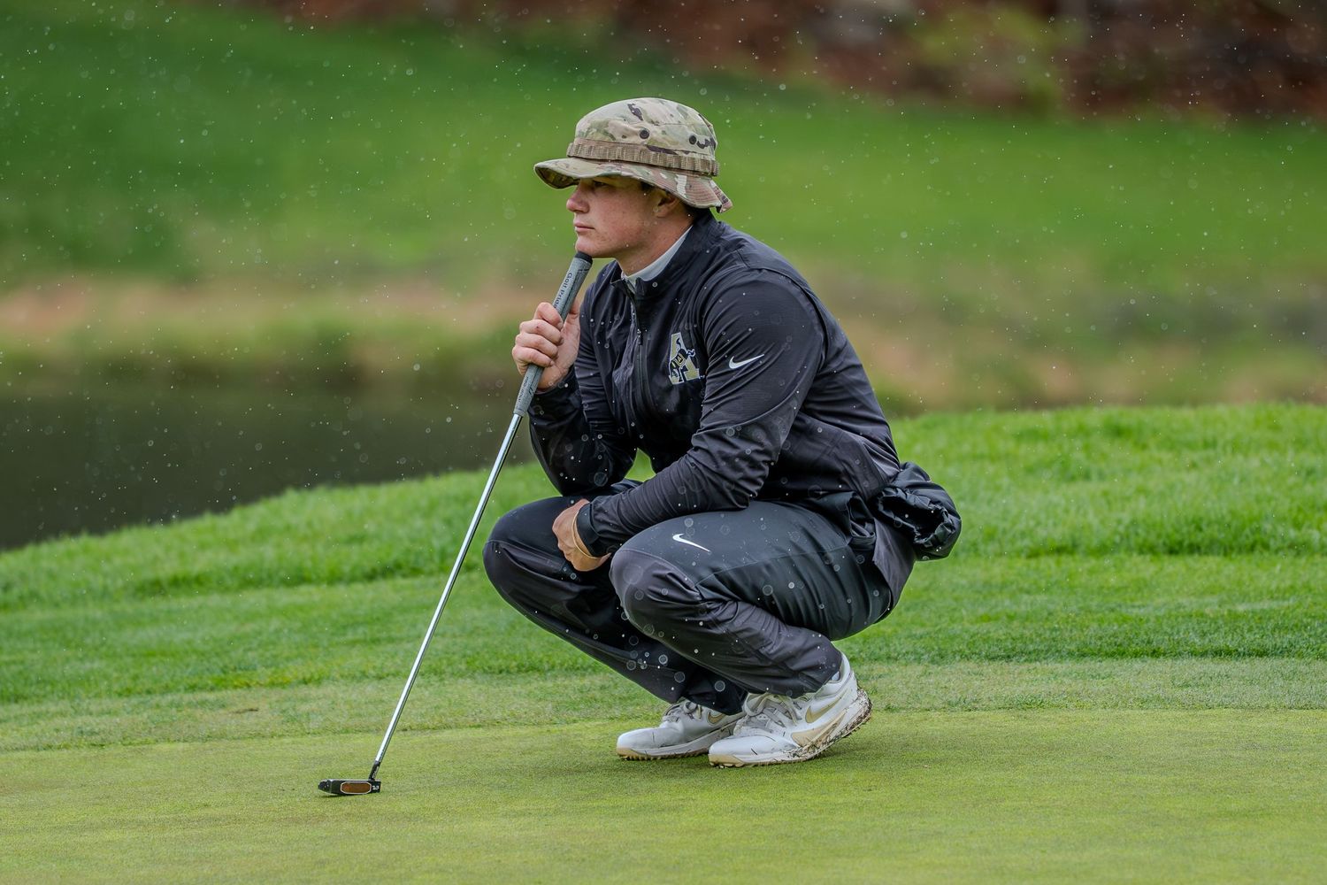 Golfer crouching on green in rain gear, analyzing putt line while holding putter on a lush golf course.