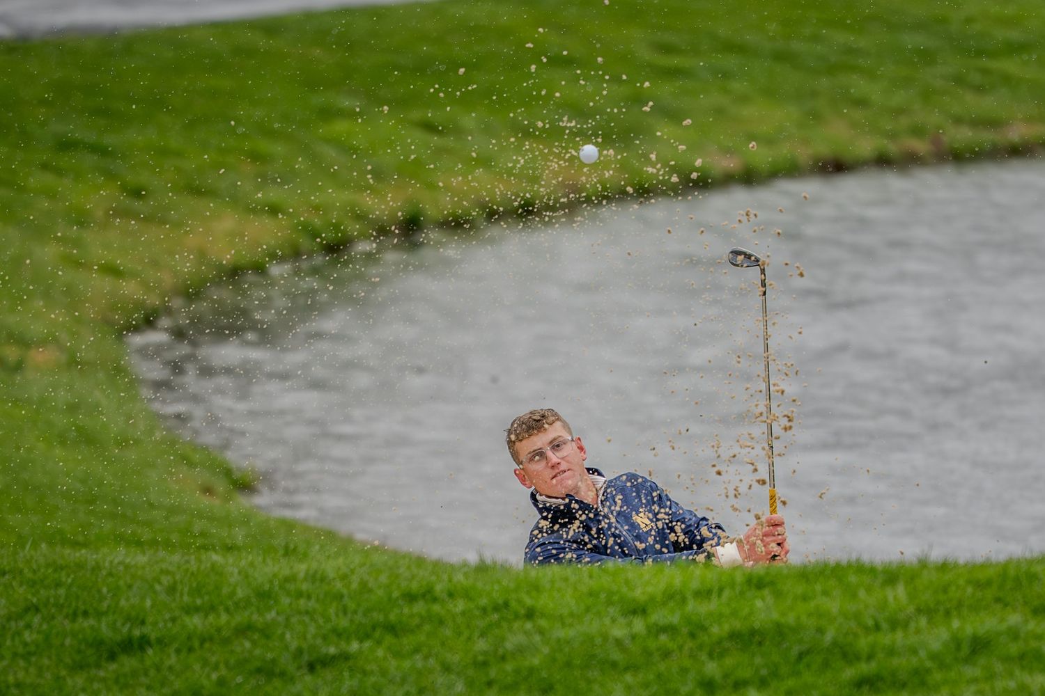 Golfer hitting a shot from a sand bunker near a water hazard on a lush green golf course.