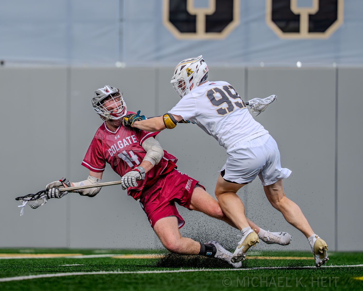 A lacrosse player in white uniform reaches for the ball while being defended.