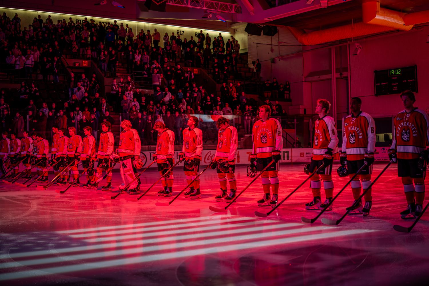 Hockey players in red and white jerseys line up on ice during a pre-game ceremony with dramatic red lighting.