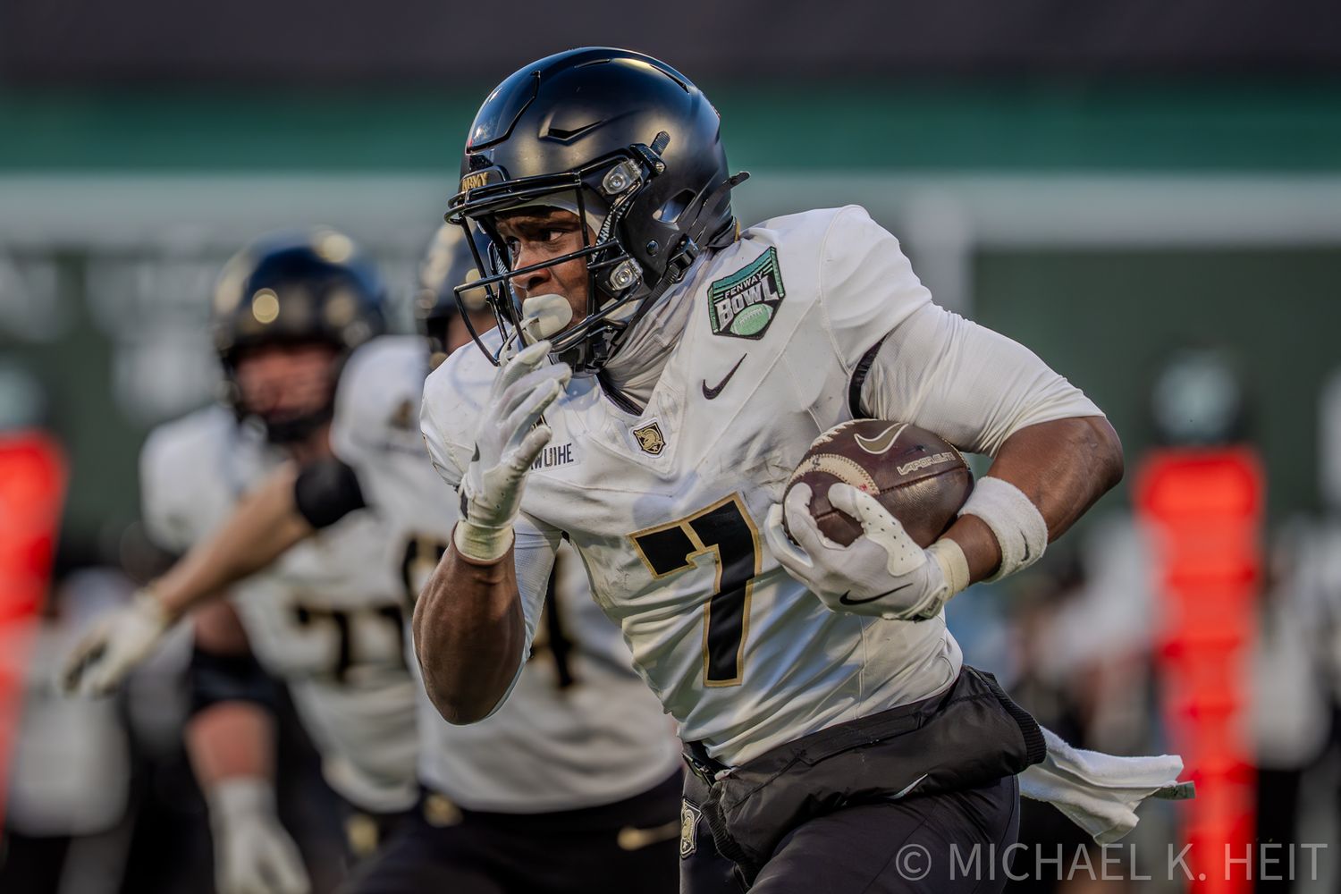 Football players in gold uniforms compete during a game on a turf field at night under stadium lights.