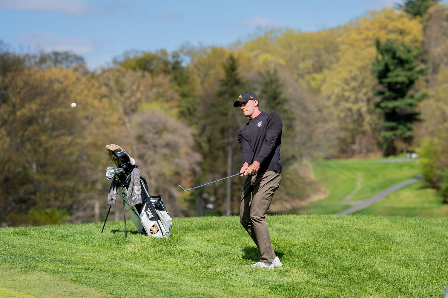 Golfer swinging a club on a lush green fairway with a golf bag nearby and trees in the background.