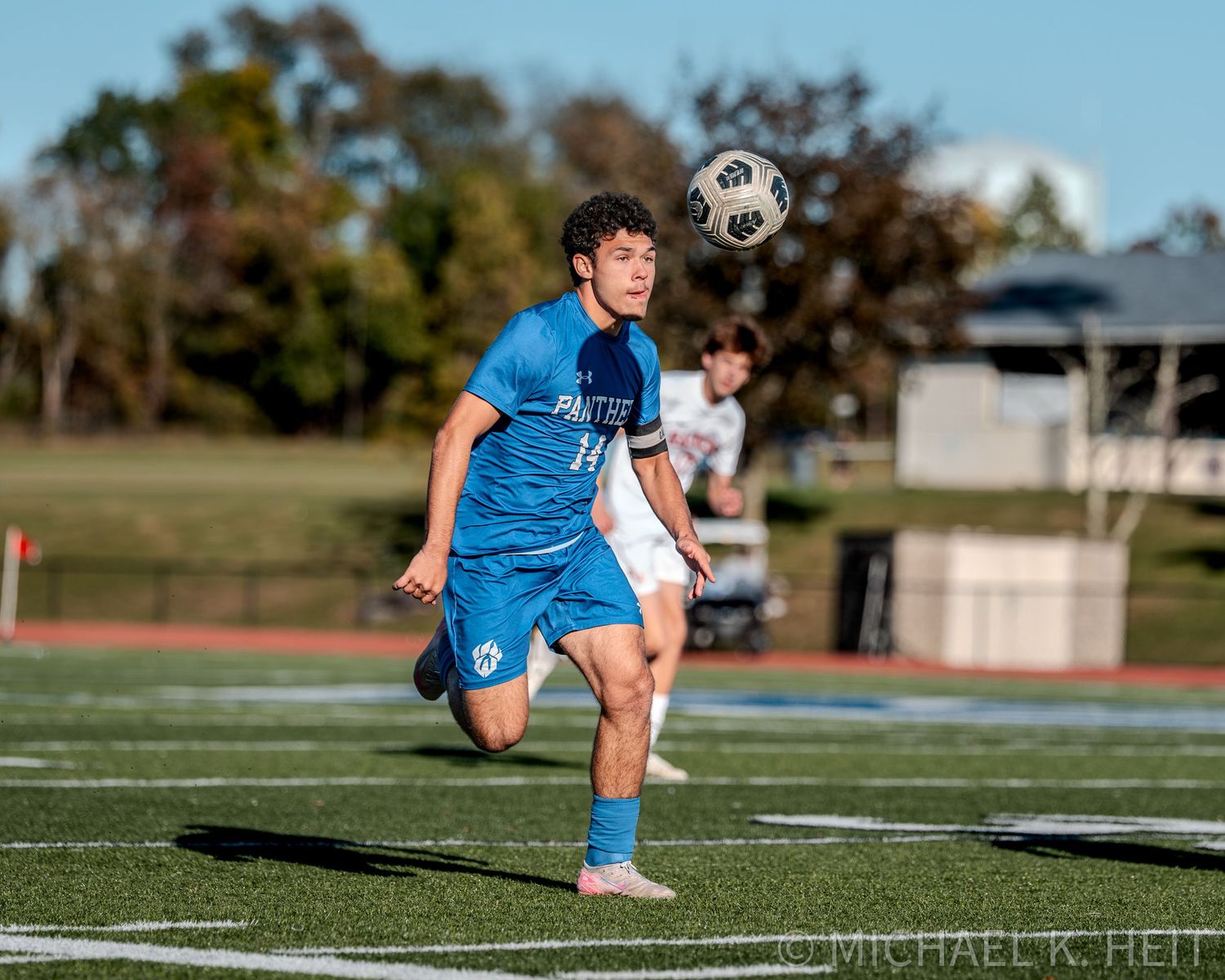 Soccer player in blue uniform stretches on field during warmup exercises in sunny outdoor setting.