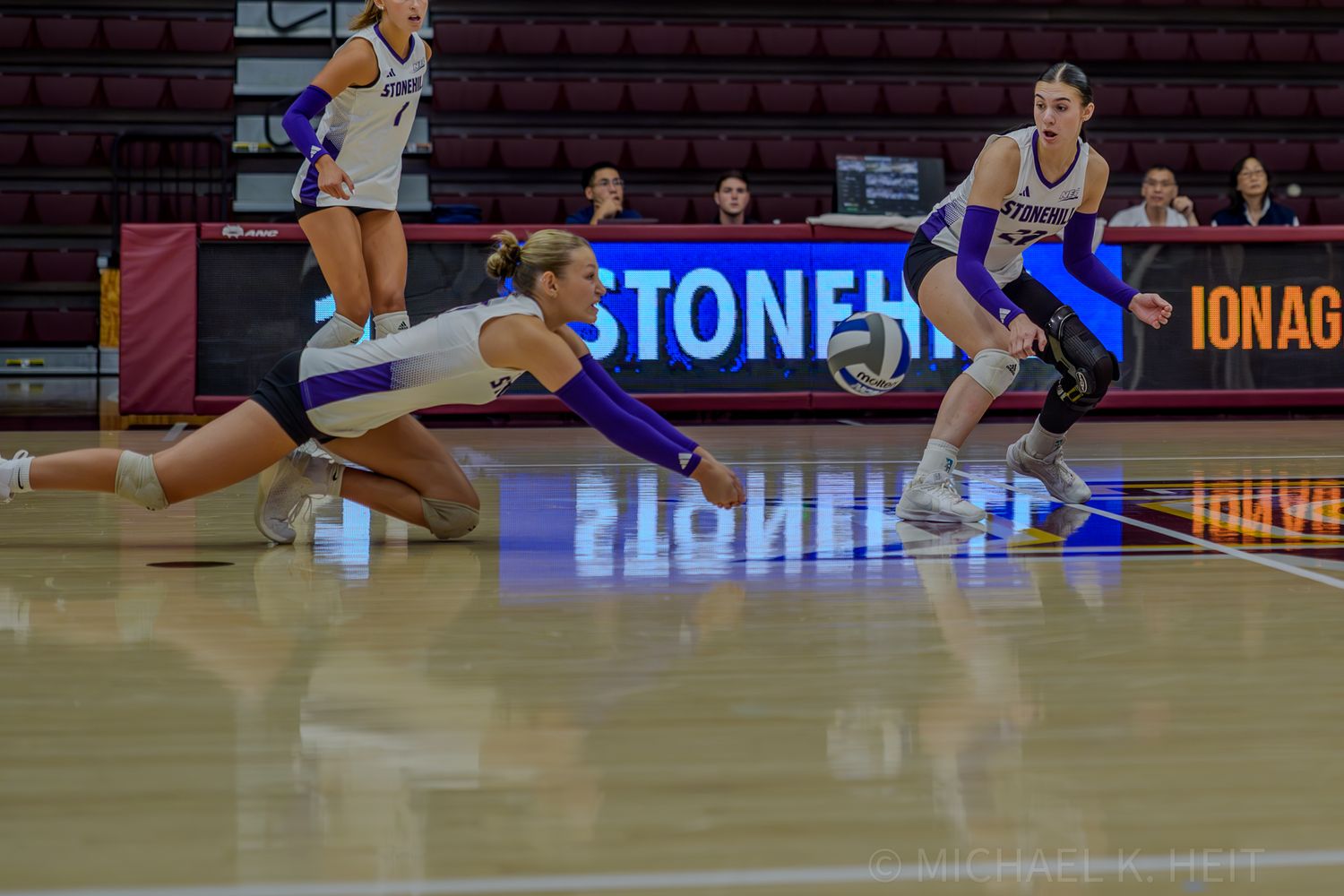 Indoor volleyball game showing players positioned for defensive play on a well-lit court.