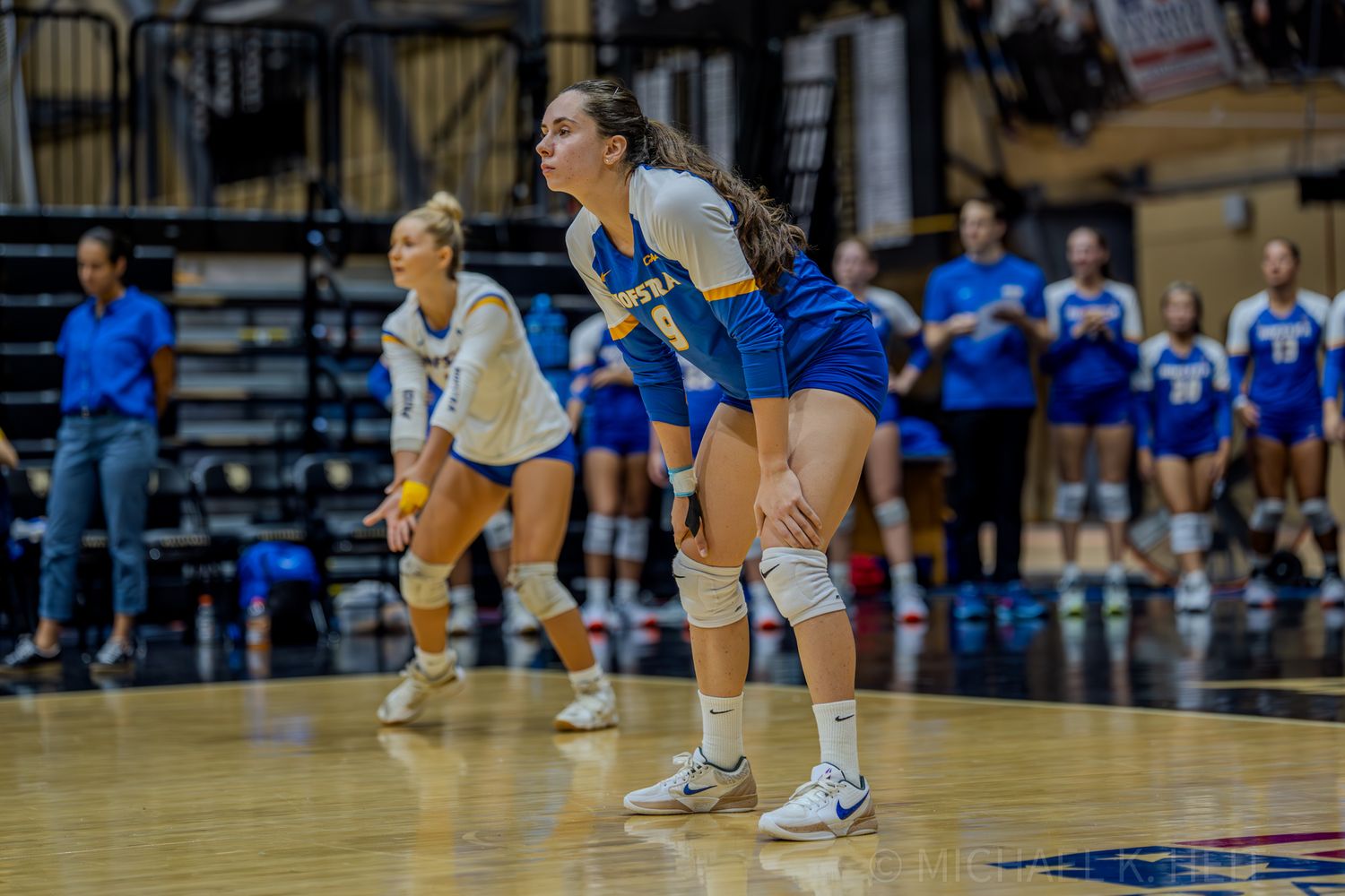 Volleyball players in blue and white uniforms position themselves defensively on the court.