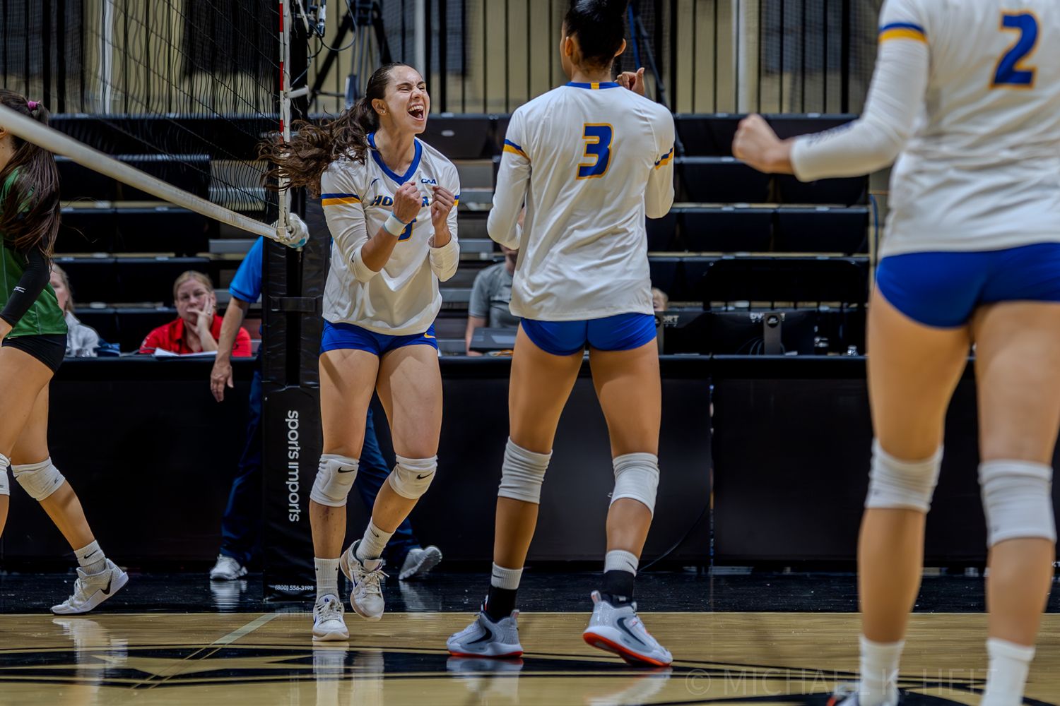 Volleyball players in blue and white uniforms position themselves on the court during an intense match.