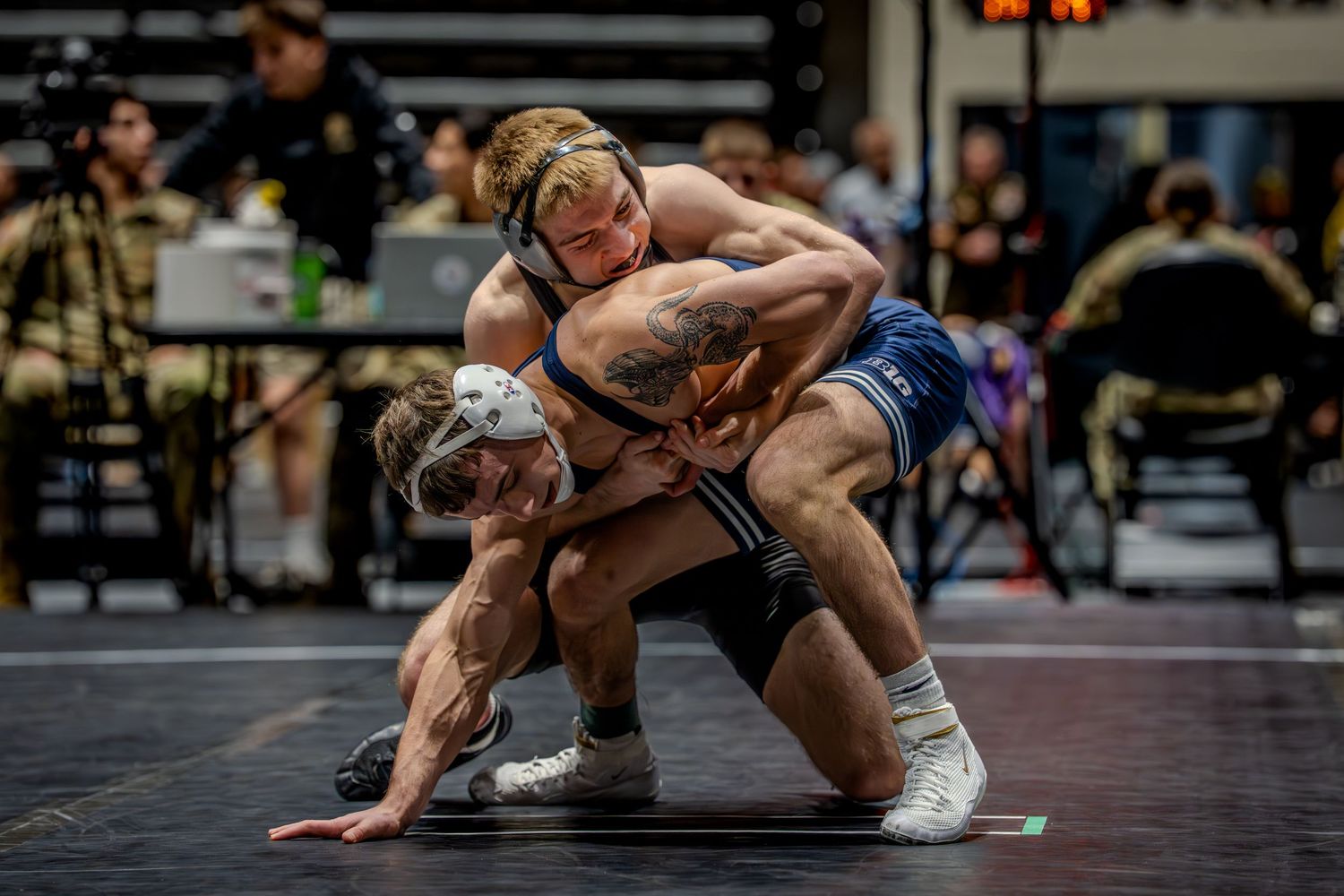 Two wrestlers compete intensely on a mat during a college wrestling match, one athlete executing a takedown move.