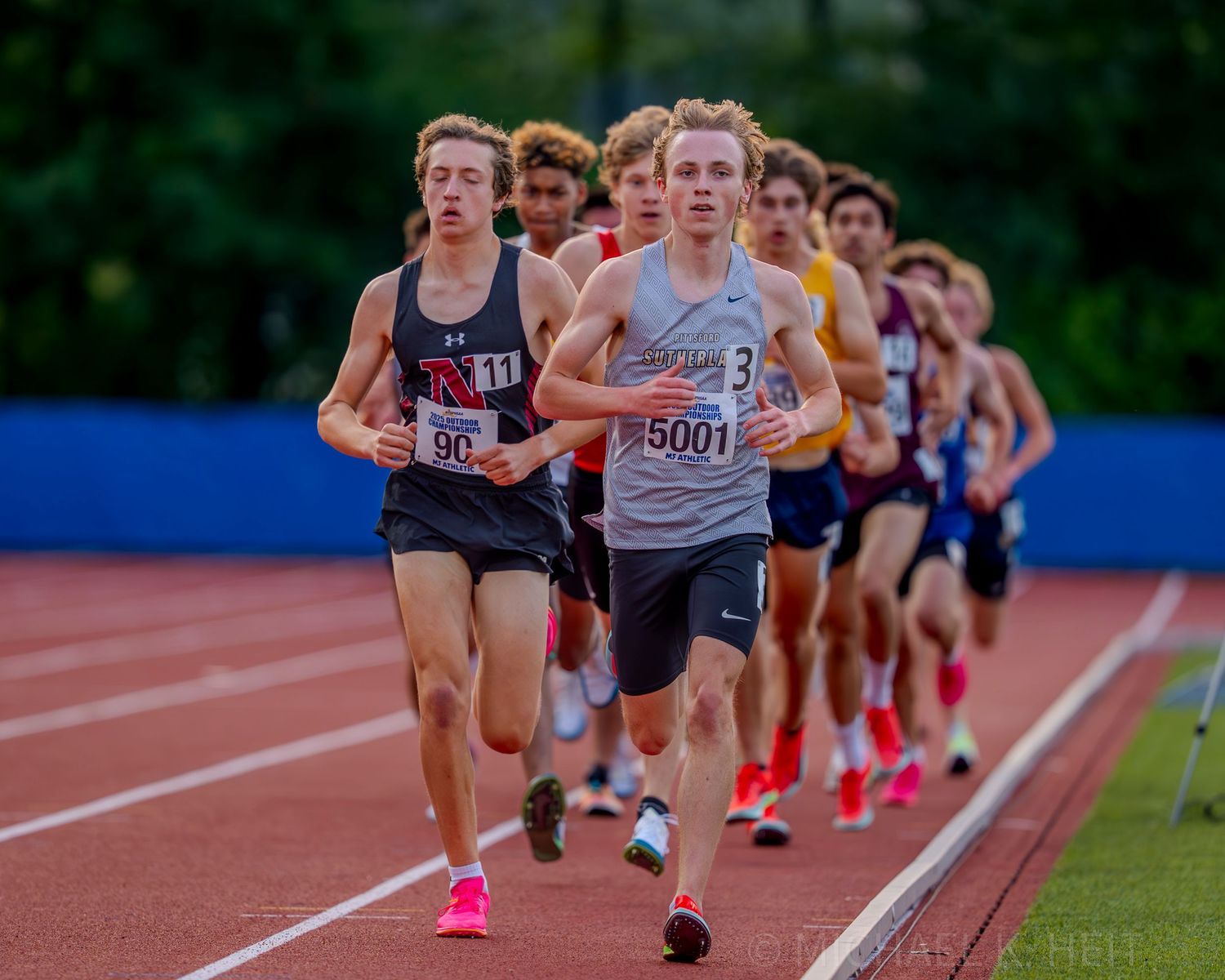 Distance runners compete in a pack during a track meet on an outdoor track.