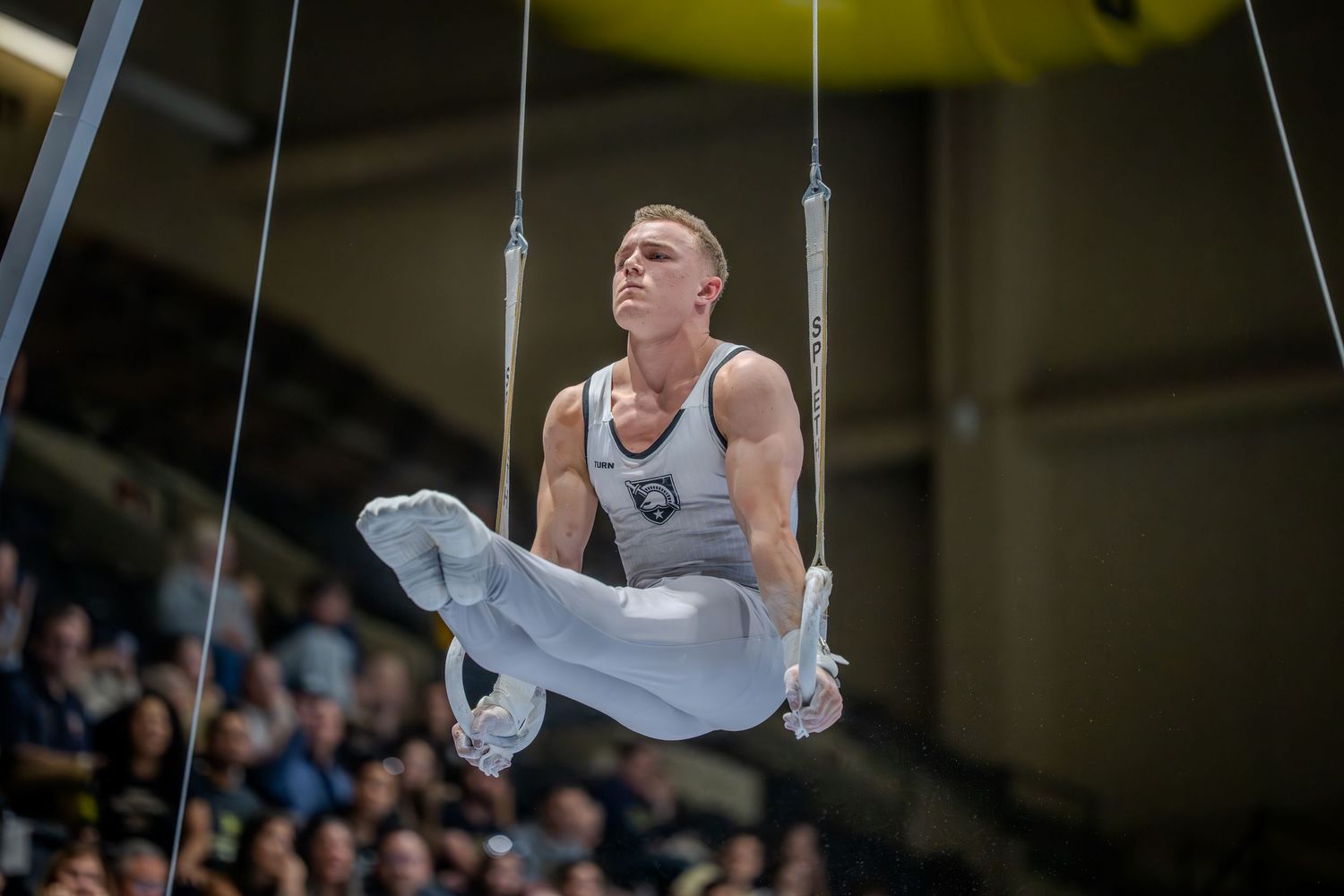 Male gymnast performing on still rings at a gymnastics competition in a packed arena.