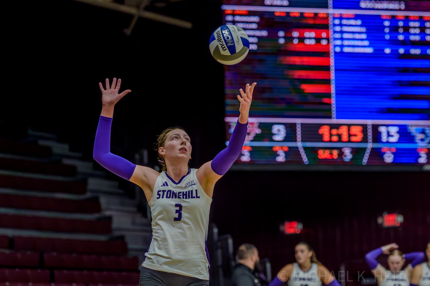 Volleyball players leap and stretch to block a shot at the net during an indoor match.