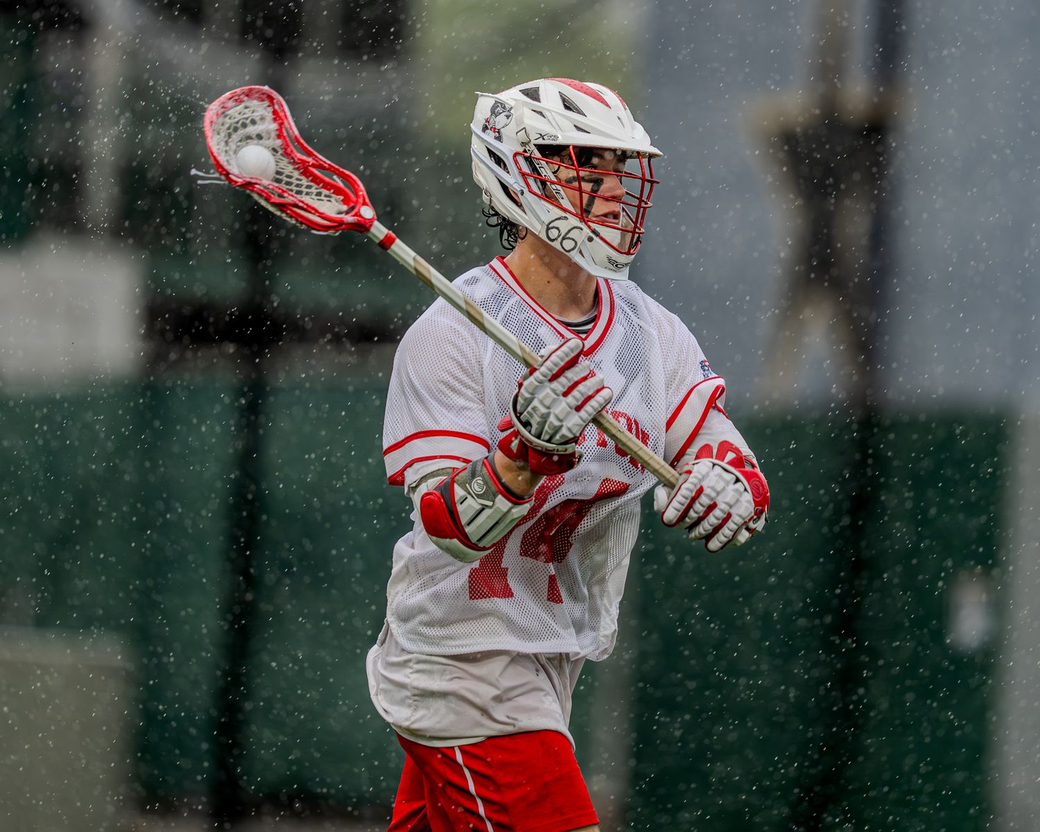 Lacrosse players in white jerseys position themselves strategically during rainy game conditions.
