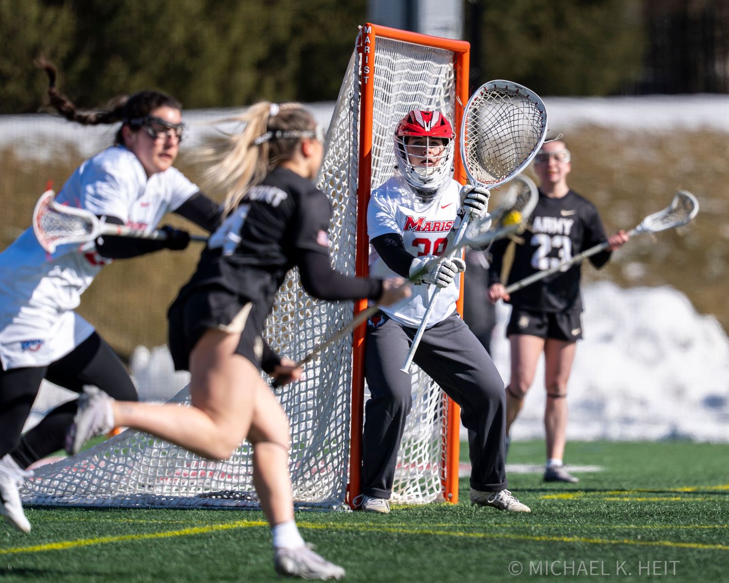 A lacrosse goalie makes a diving save in front of the net during a match on a sunny day.