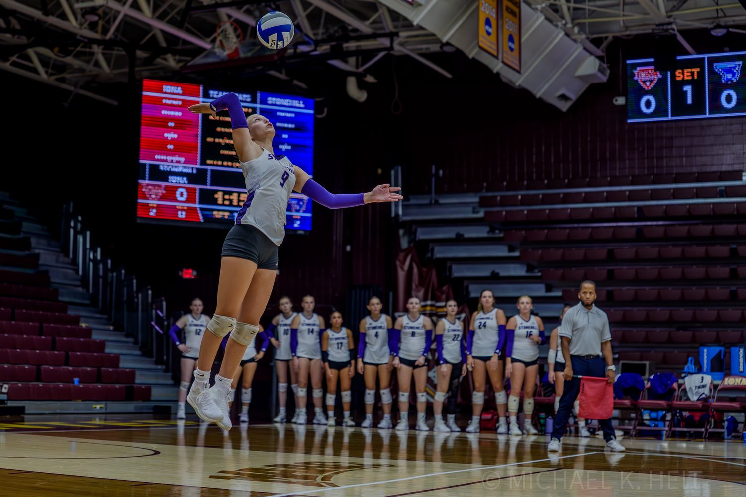 Volleyball player leaps high for a spike while teammates watch from the sideline.
