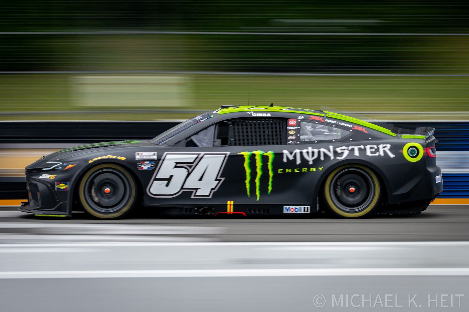 NASCAR Monster Energy #54 race car speeding on track, captured with motion blur effect against green trees backdrop.