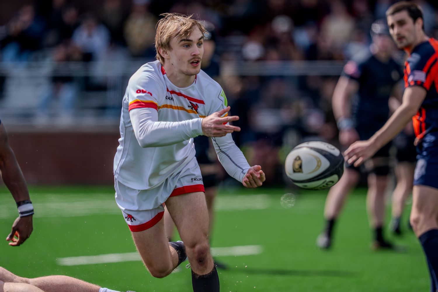 Rugby player in white uniform passes the ball during an intense match against opponents in dark jerseys.