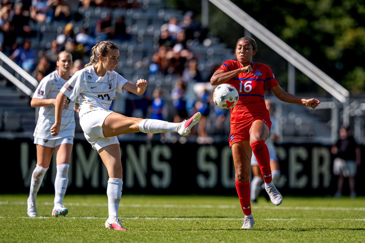 Two female soccer players compete for the ball during a collegiate match, one in white and one in red uniforms.