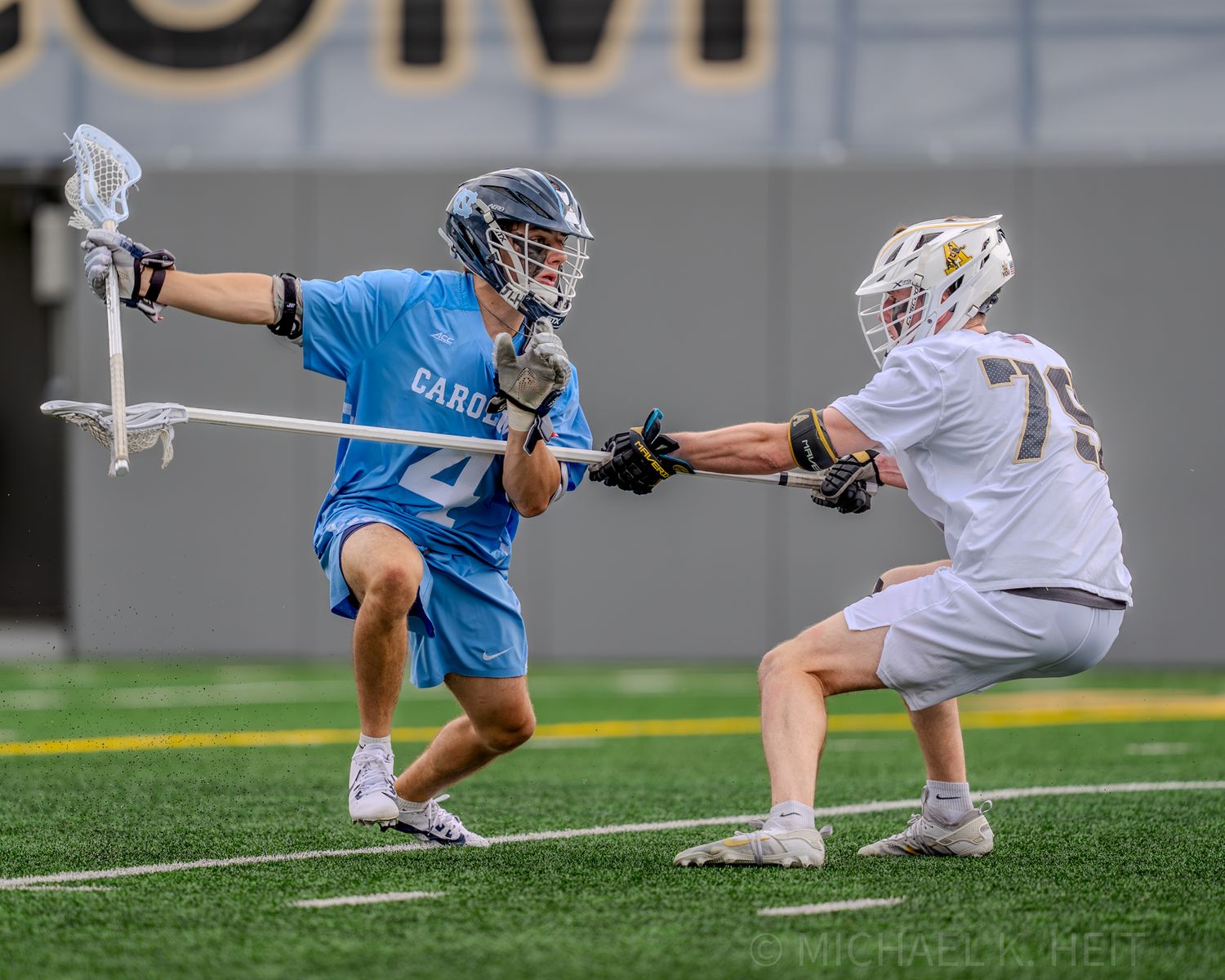Lacrosse players in white and light blue uniforms competing for ball possession.