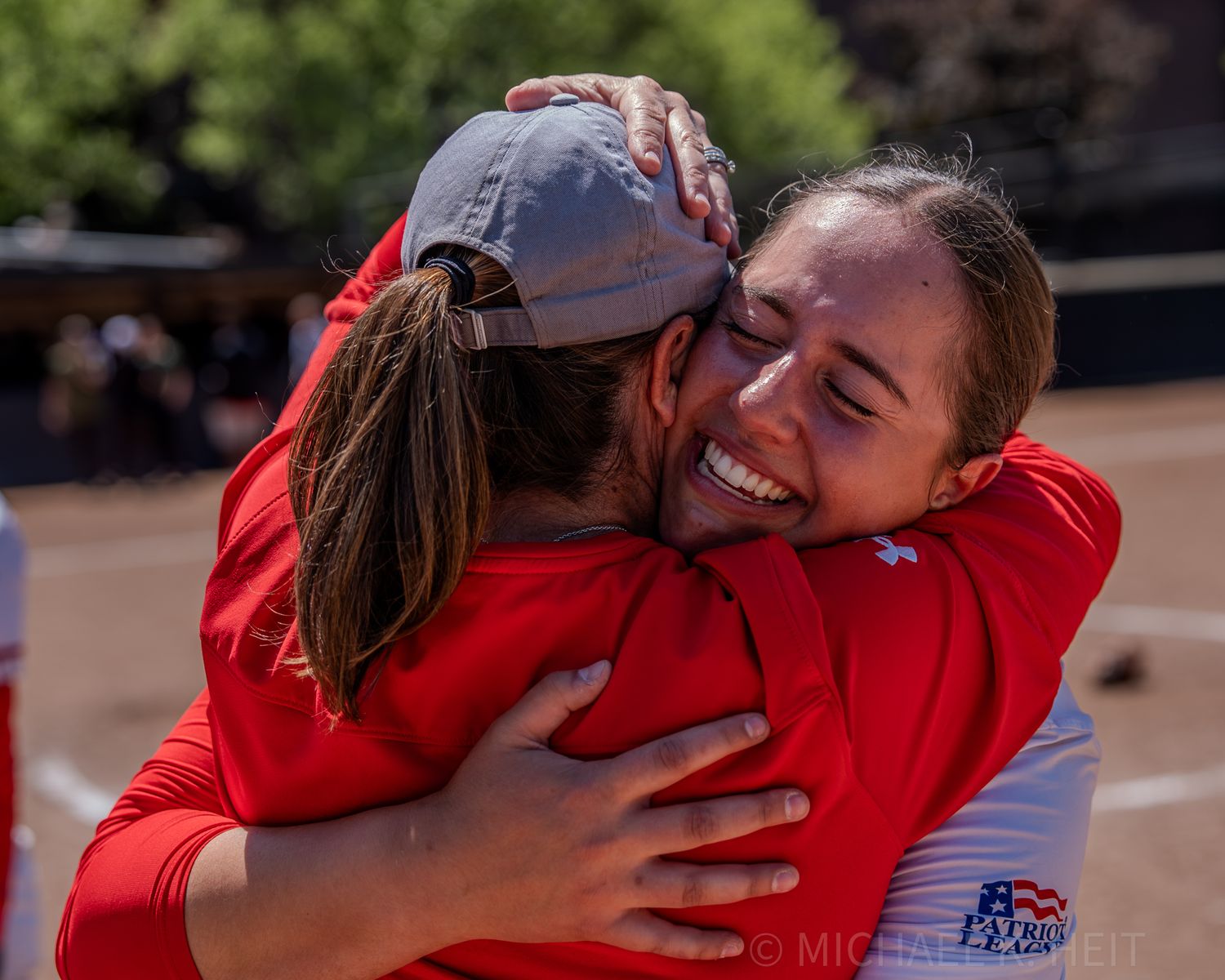 Athletes sharing emotional embrace in red uniforms on softball field.