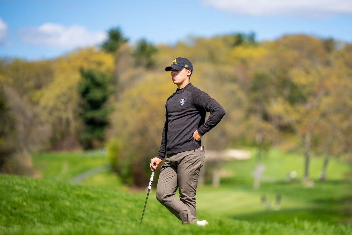 Golfer in black outfit standing on lush green golf course with scenic tree-lined background on a sunny day.