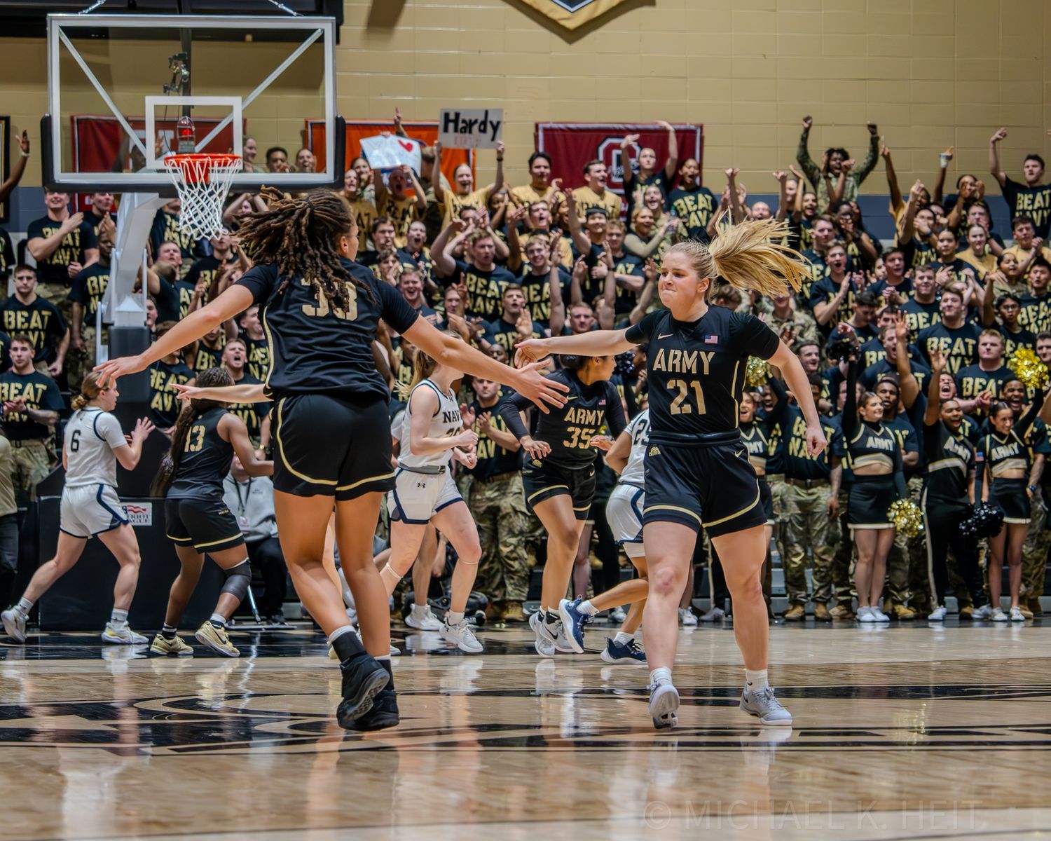 Basketball players compete on a wooden court with military personnel and fans watching from the sidelines.