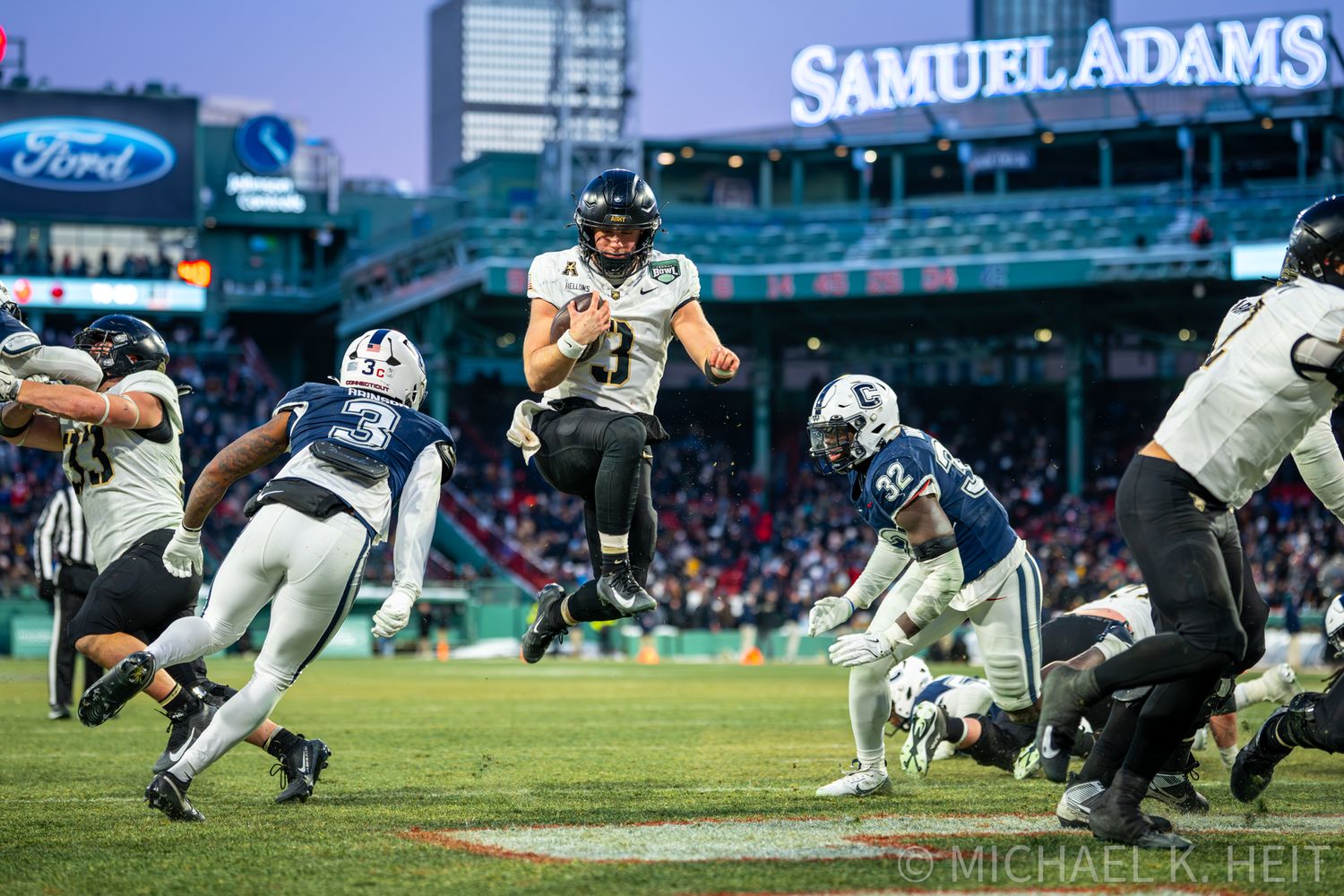Football player leaps high into the air during a game at a stadium with Samuel Adams signage visible.