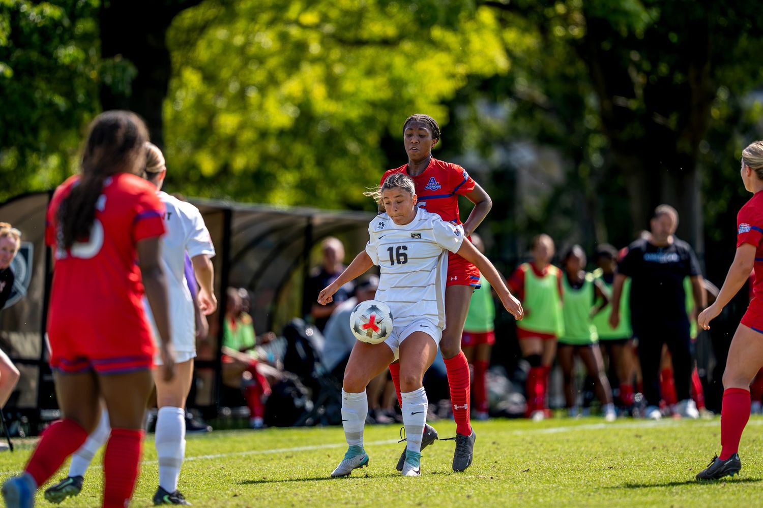 Female soccer player #16 in white uniform controls the ball during a college soccer match on a green field.