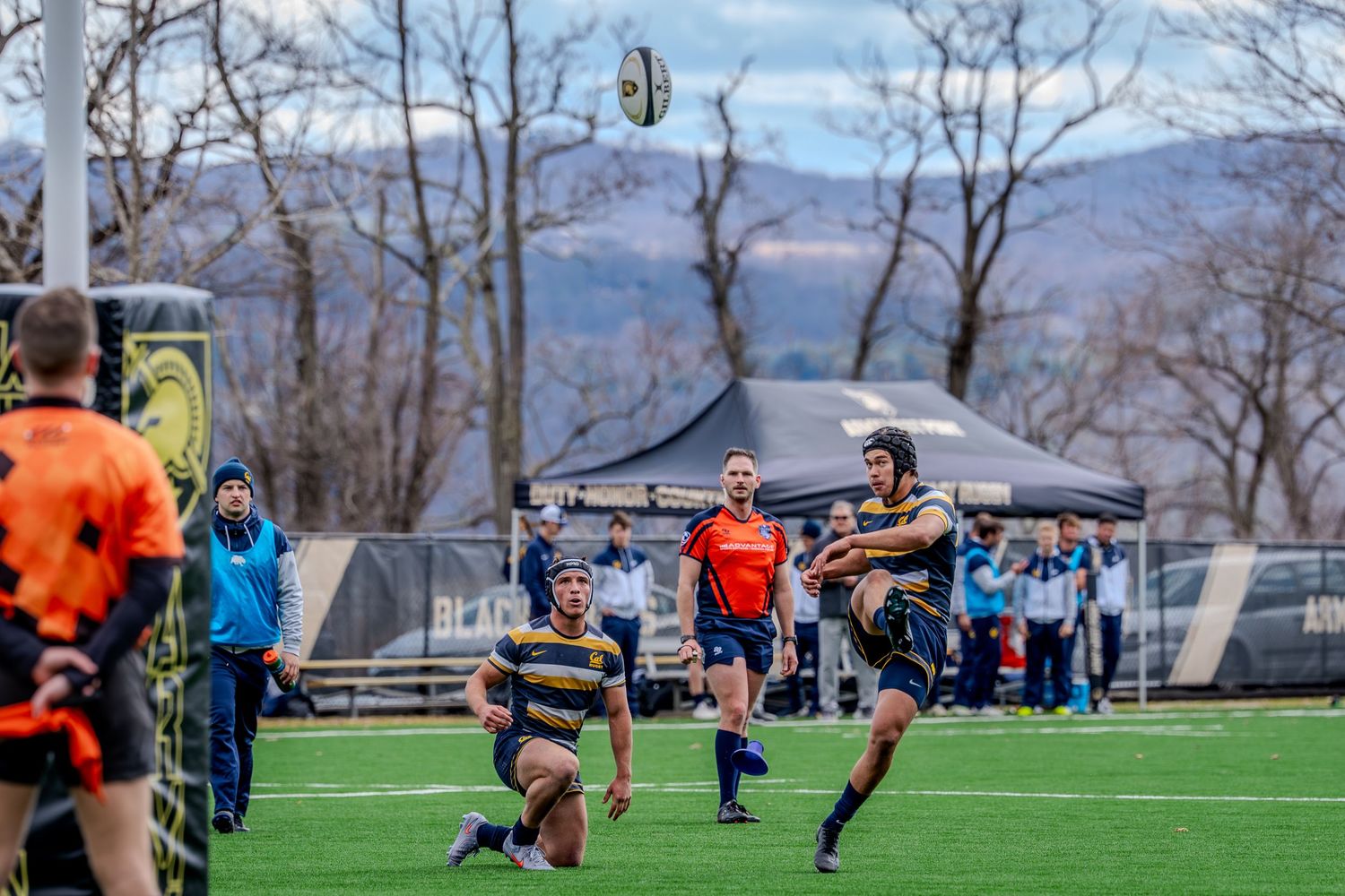 Rugby players compete in a match on an artificial turf field with mountains and bare trees visible in the background.