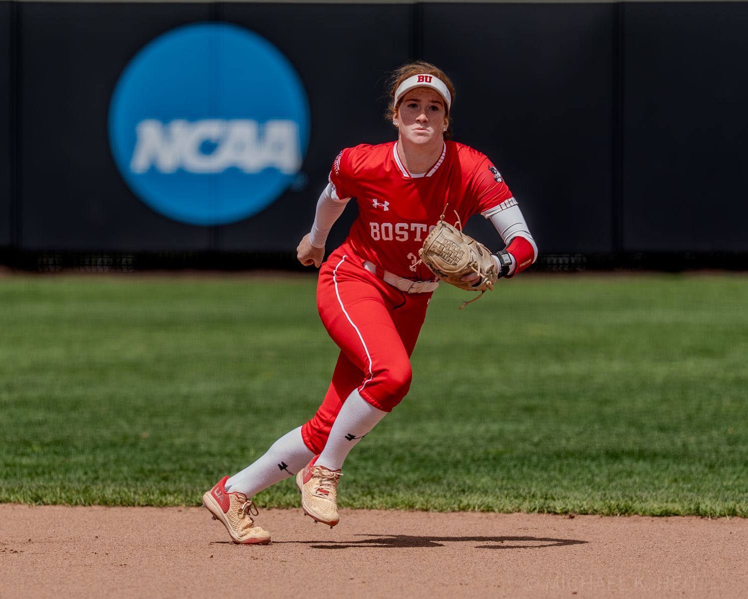 Baseball player in red uniform crouches in defensive position at shortstop during NCAA game.