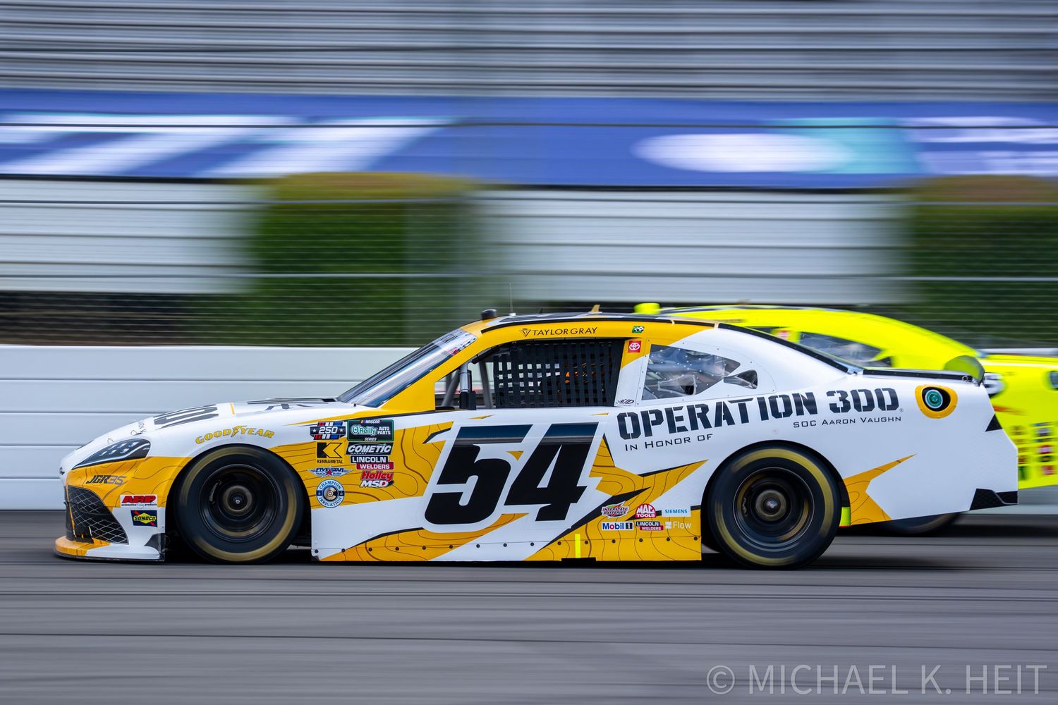 NASCAR race car #54 Operation 300 yellow and white livery speeding on track during a race event.