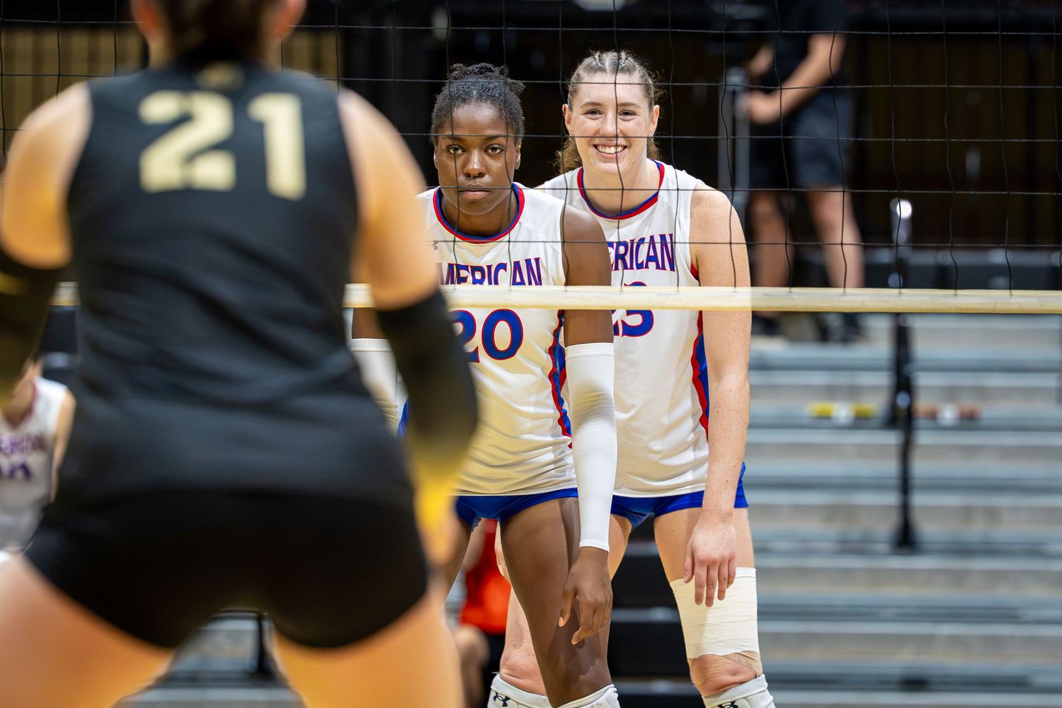 Two American University volleyball players in white uniforms stand at the net during a competitive indoor match.