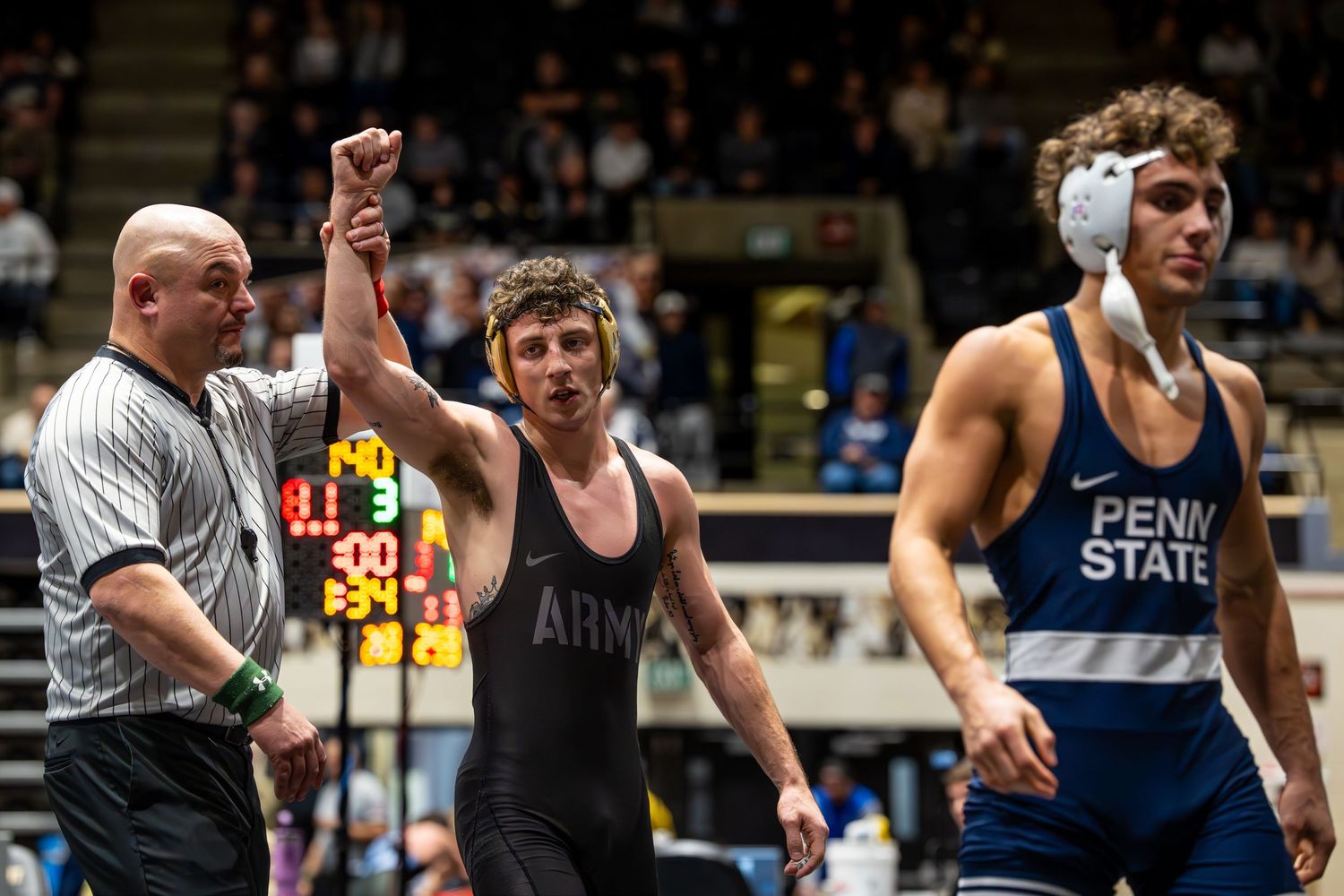 Army wrestler celebrates victory with referee raising hand, facing Penn State opponent on wrestling mat.