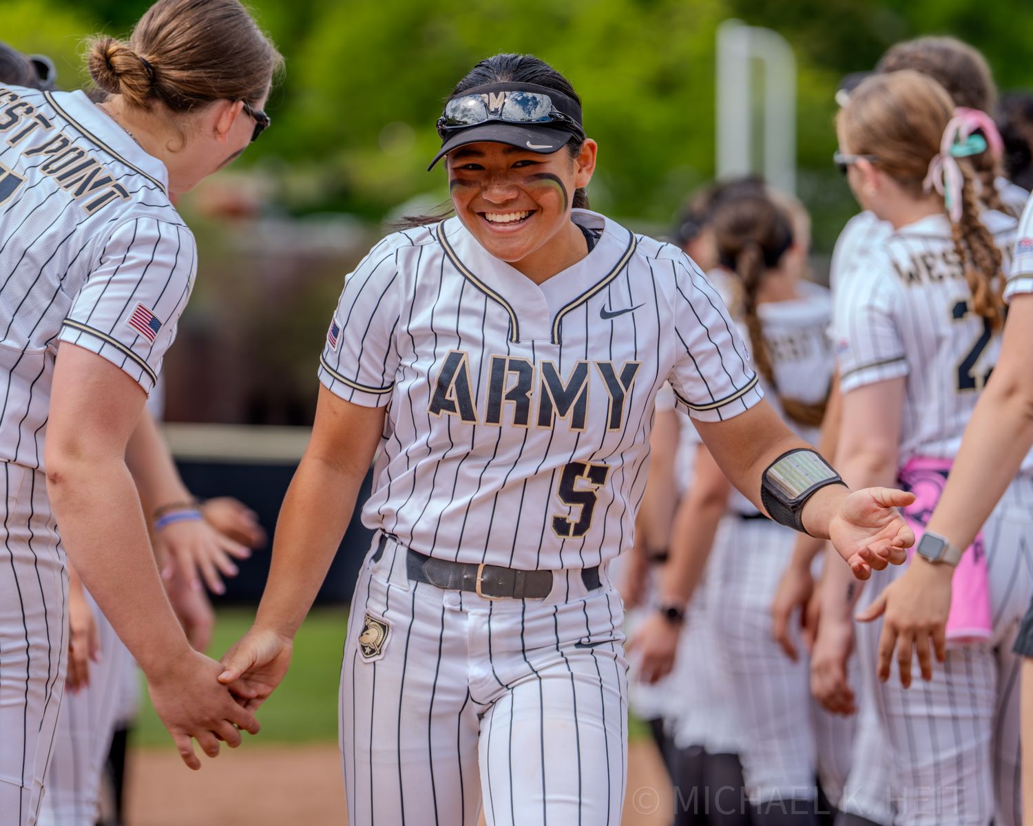 Army softball players in pinstriped uniforms interact along dugout railing during game.