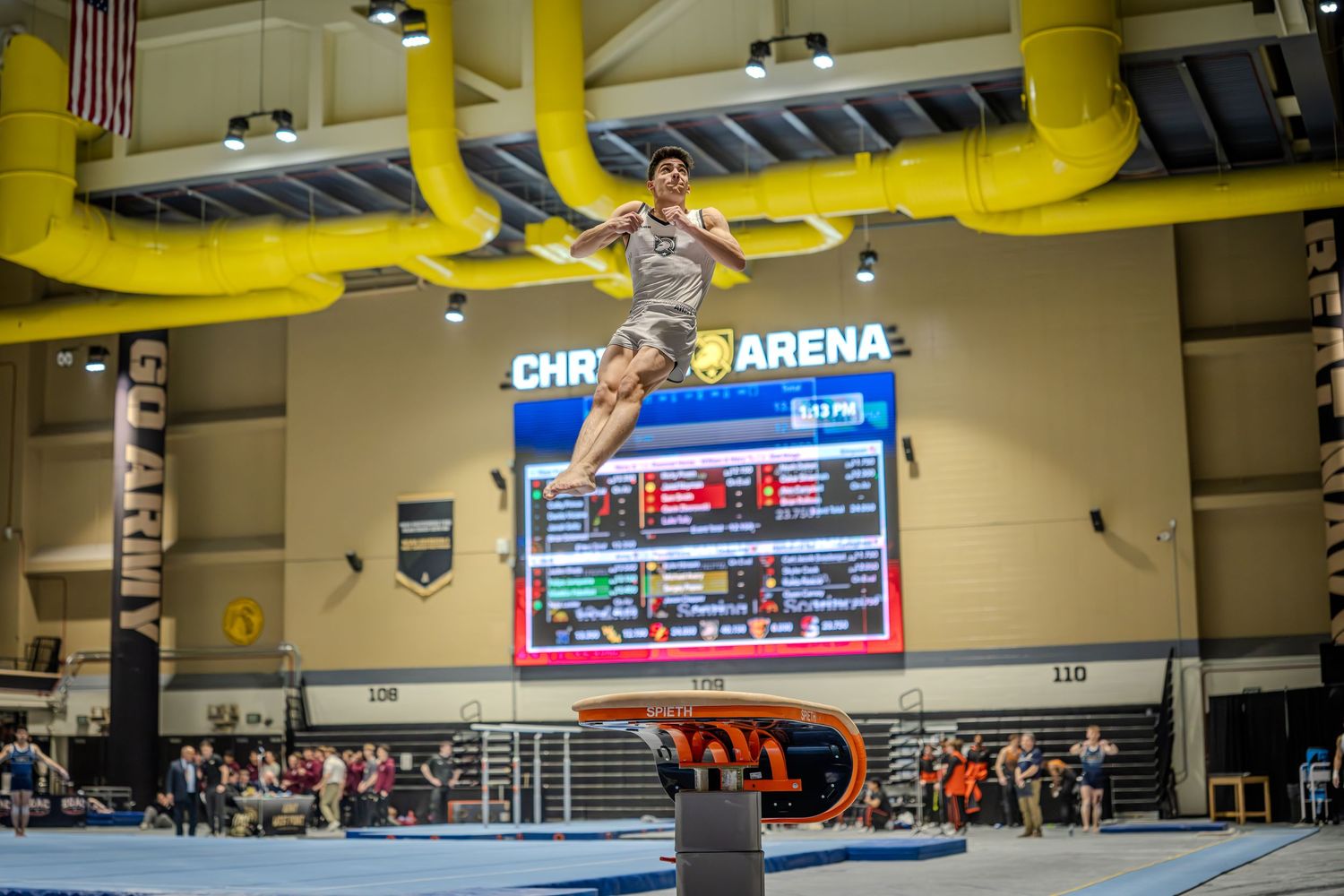 Gymnast performing a high vault jump over a pommel horse in an indoor arena with scoreboard visible.