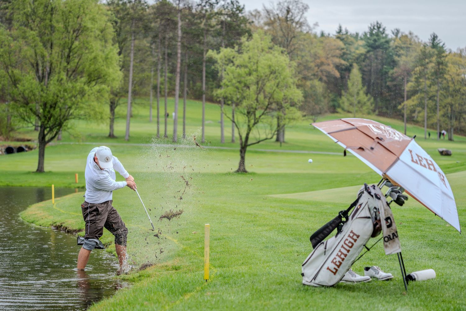 Golfer hitting a shot from a water hazard on a lush green golf course, with a golf bag nearby.