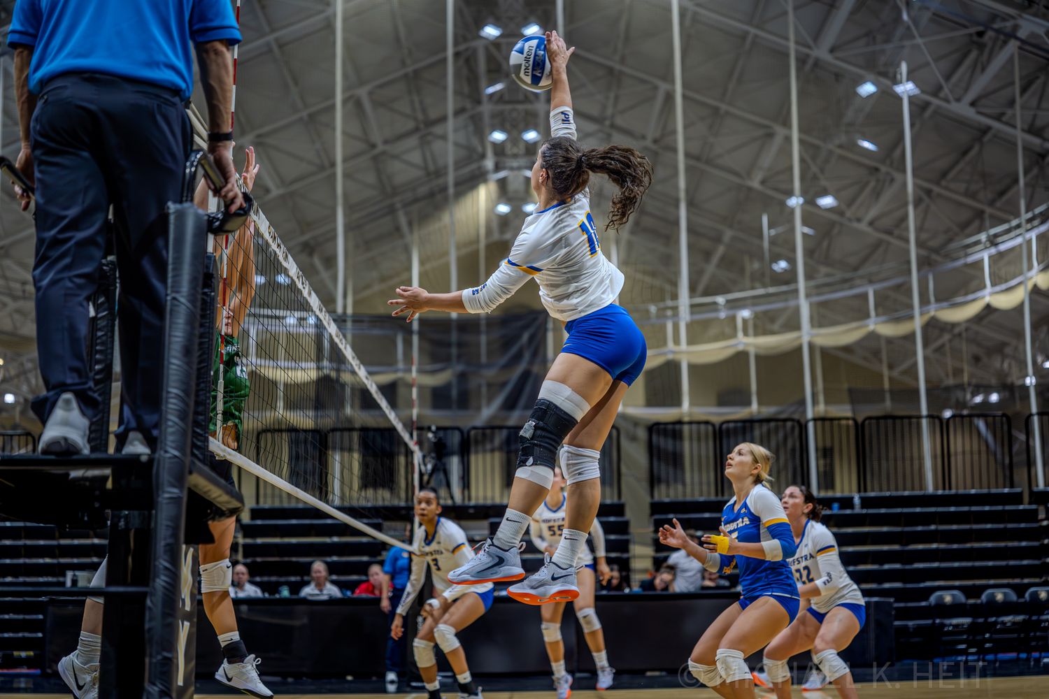 Players positioned defensively on volleyball court with high ceiling gymnasium lighting visible.